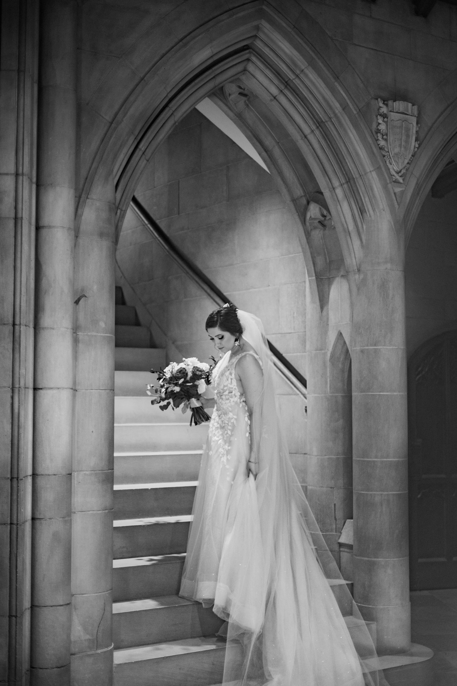 Black and white photo of bride in lace gown standing in arched church doorway.