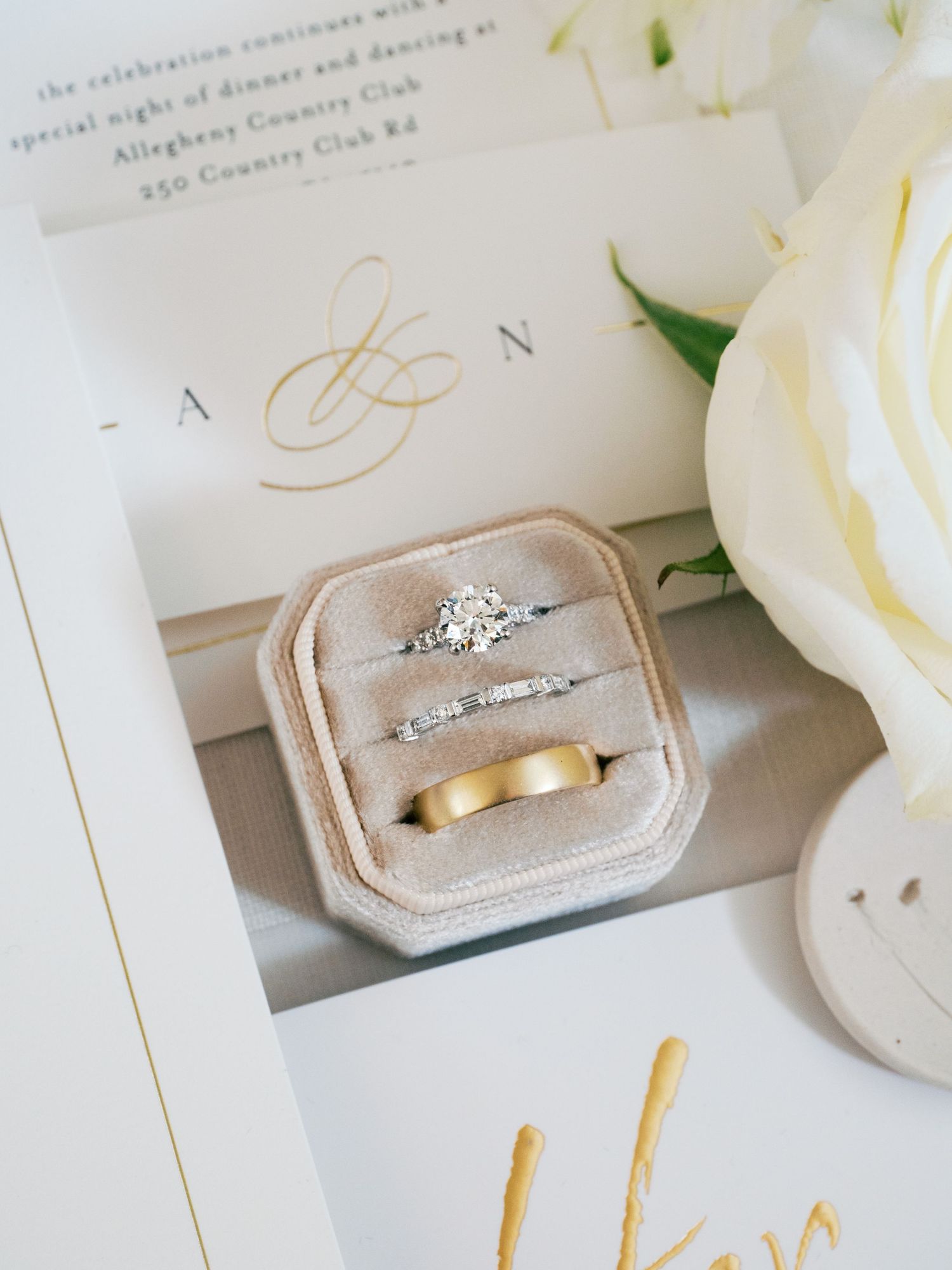Wedding rings displayed in a blush velvet ring box next to white roses and invitation details.