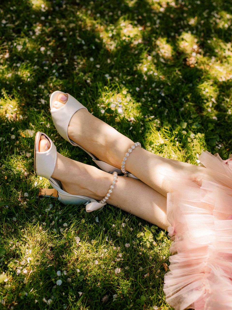 Pearl-adorned bridal shoes on feet resting on mossy ground with tulle dress visible.