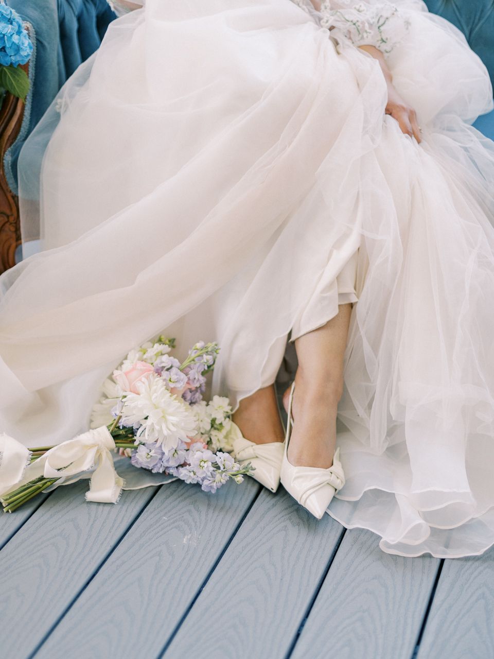 Wedding shoes and bouquet peek out from under flowing white wedding dress on blue deck.
