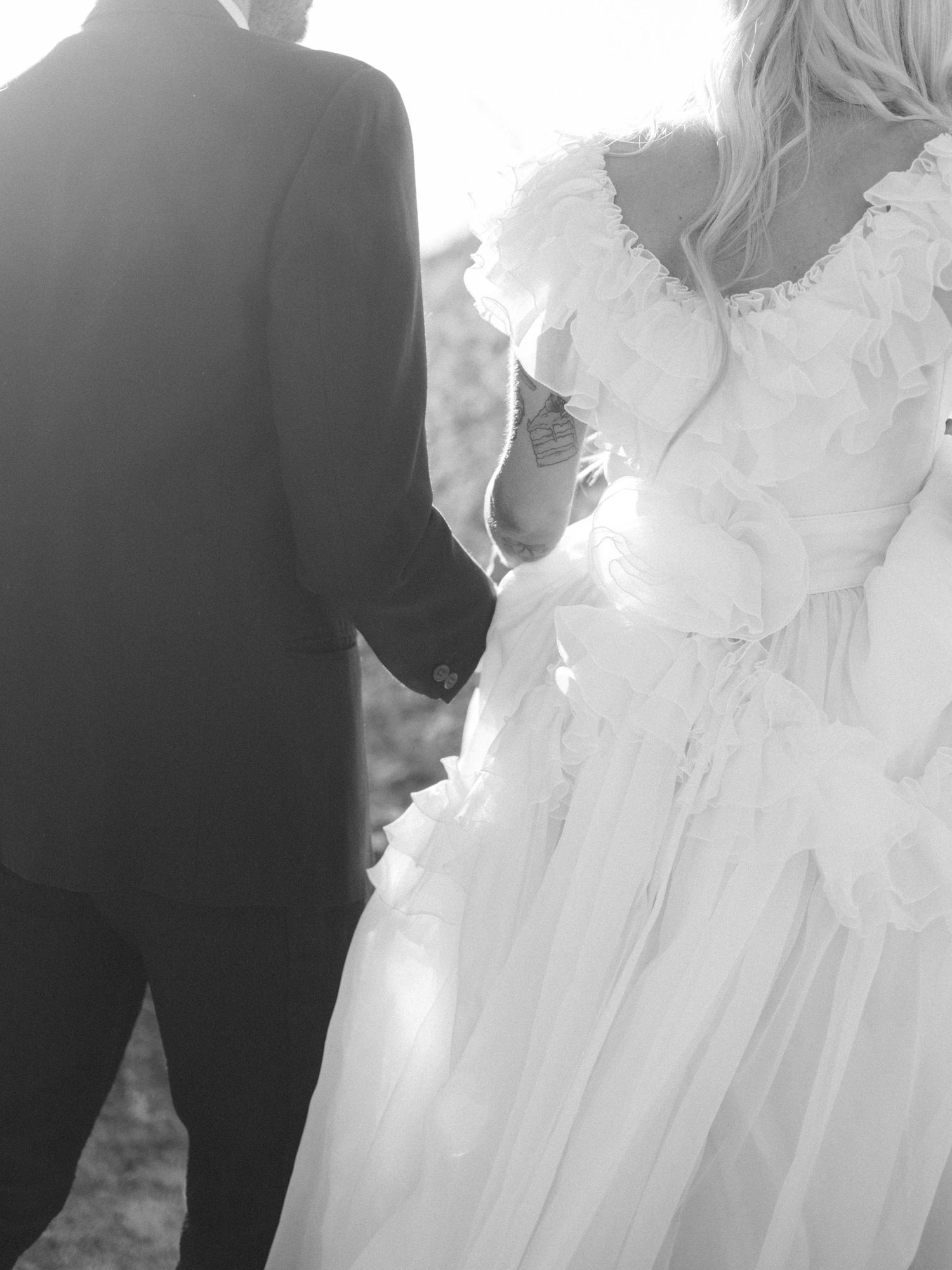 Black and white photo showing back details of a wedding dress and groom's tuxedo during an intimate moment.