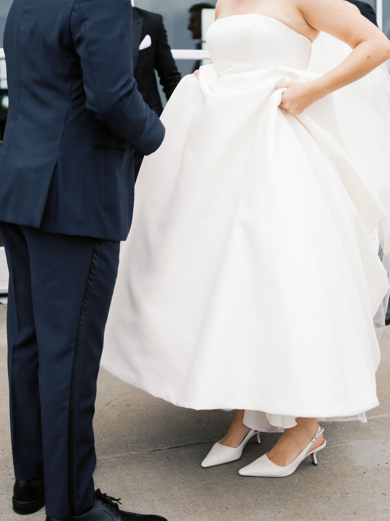 Candid wedding photo showing details of white wedding dress with groom in navy suit.