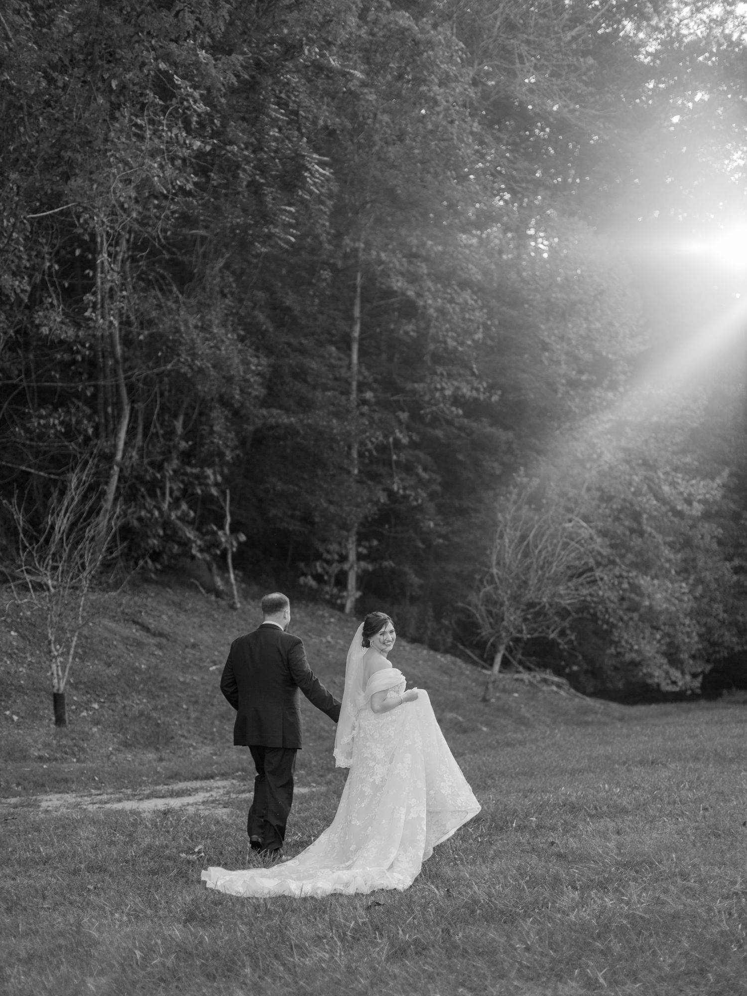 A black and white wedding photo of a couple standing in sunlit woodland.