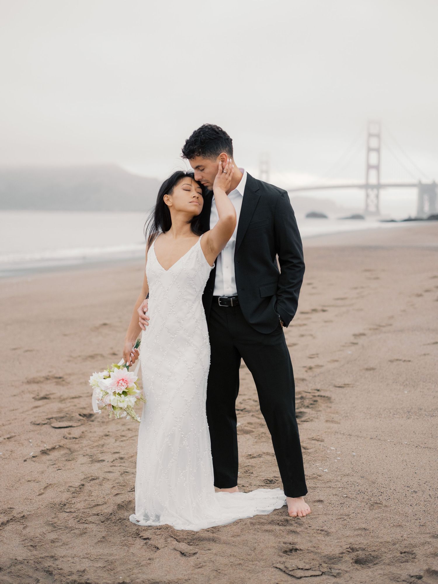 A romantic wedding couple embraces on Baker Beach with the Golden Gate Bridge visible through fog in San Francisco.