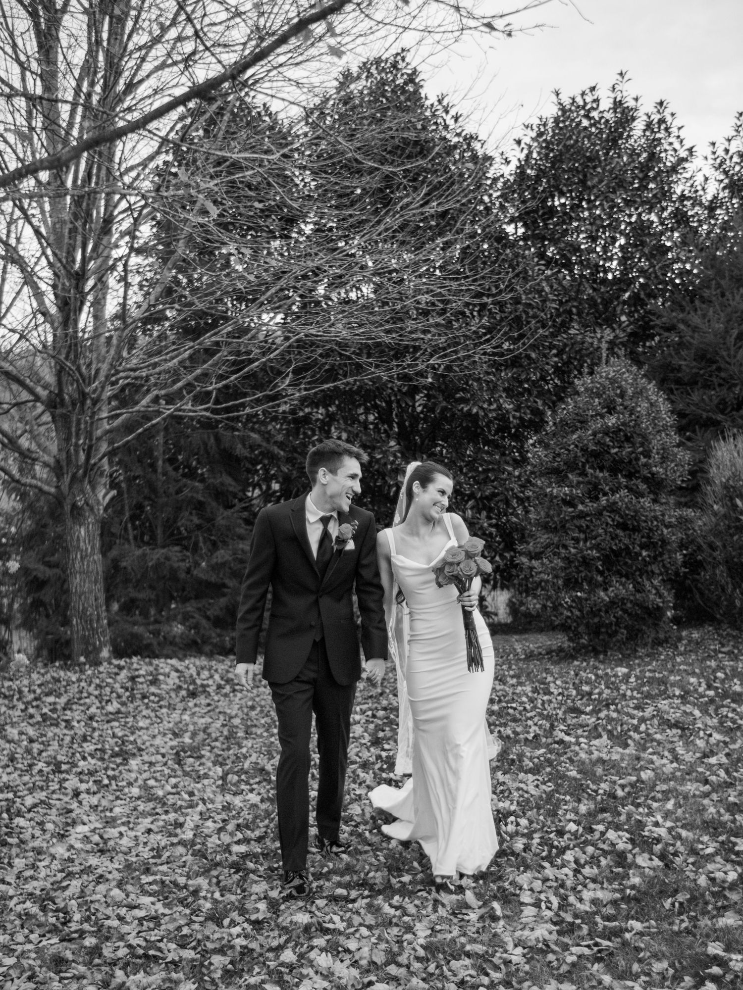 A couple in wedding attire walk together on fallen autumn leaves in a wooded setting.
