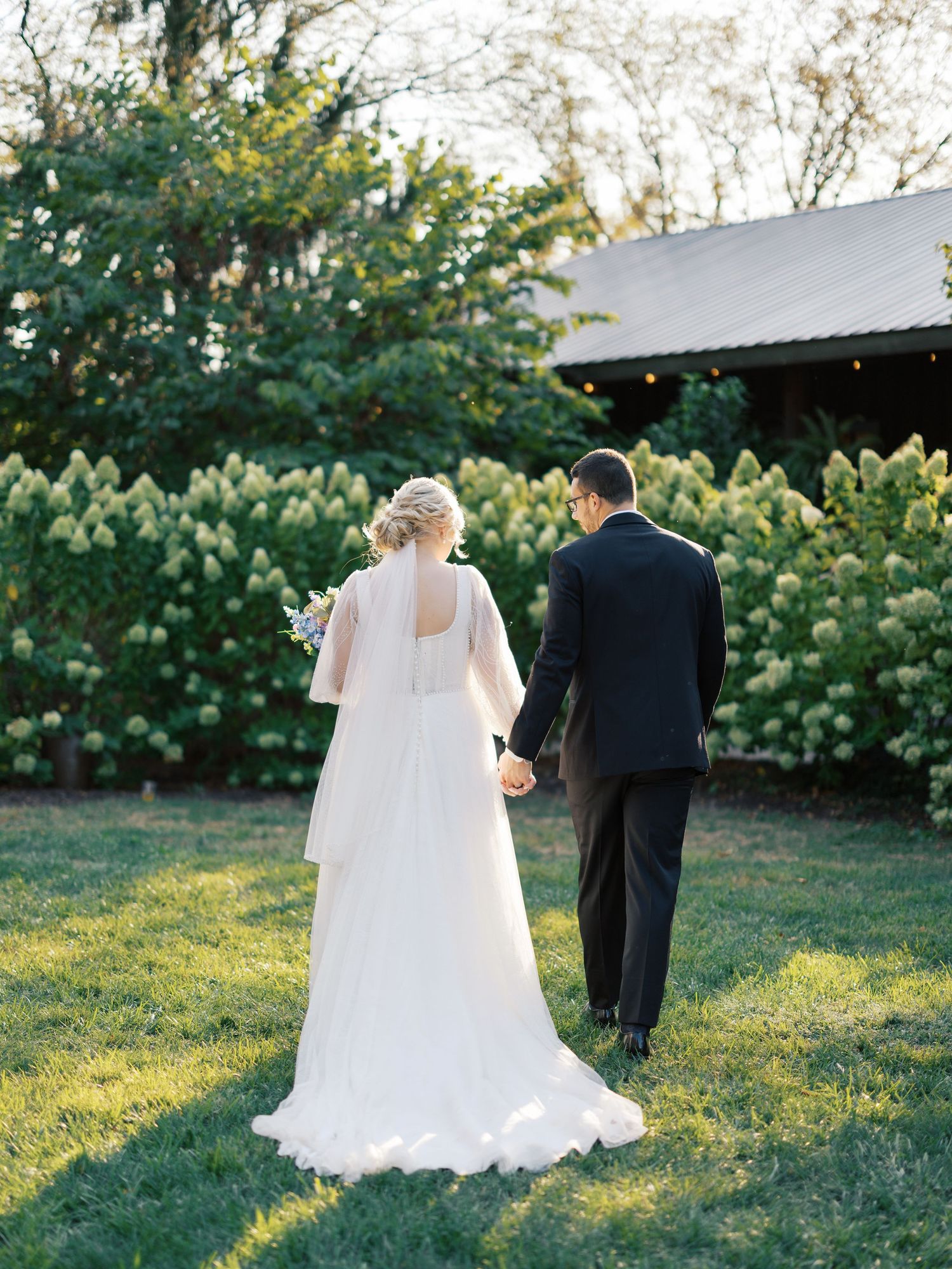A bride and groom walking on a garden lawn with flowering bushes in the background.