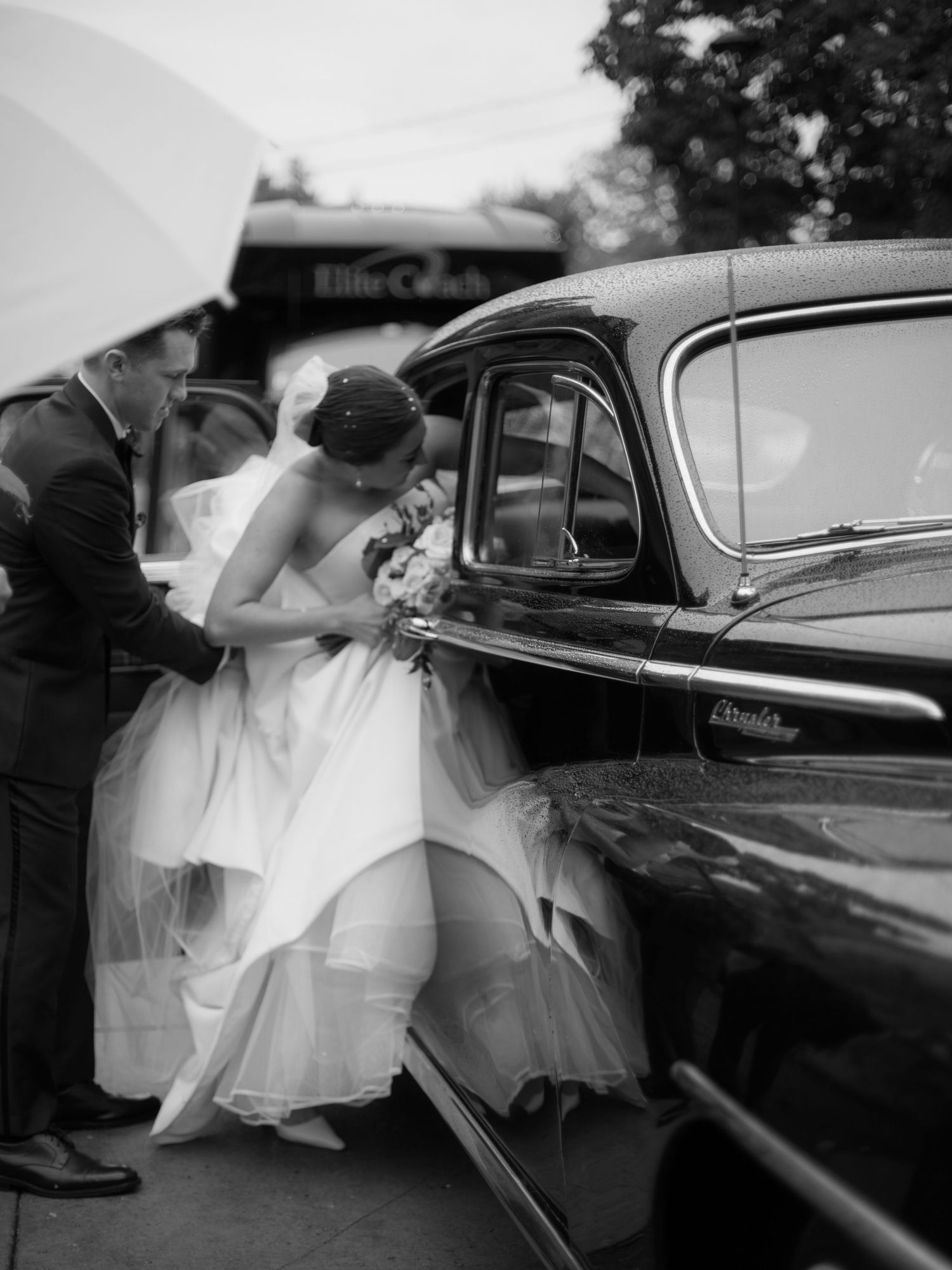 A vintage black and white photo showing a person in a fluffy wedding dress being helped into a classic car.
