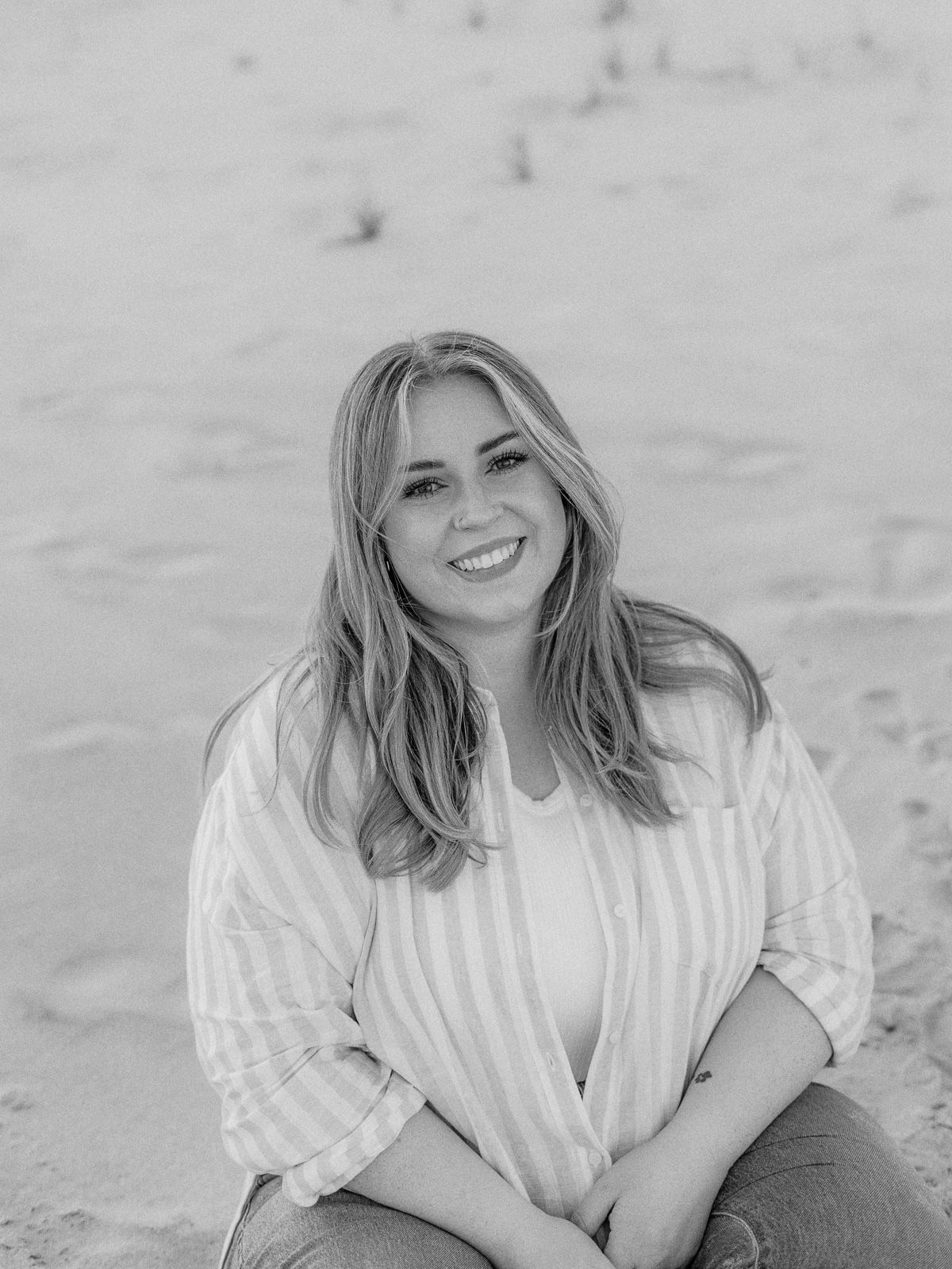 Black and white portrait of someone with long hair and a white blouse sitting on a beach, smiling warmly at the camera.