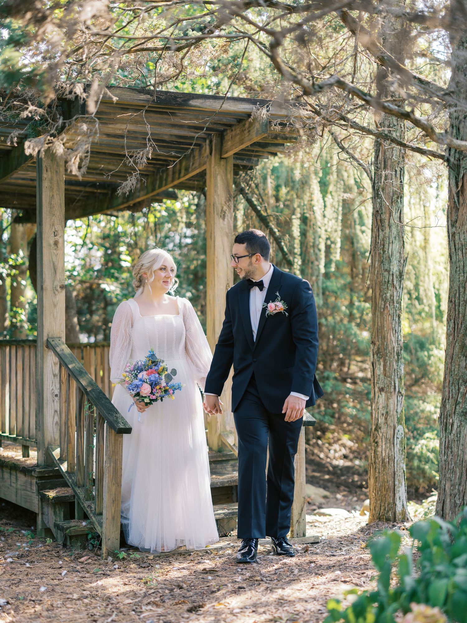 A couple stands under a wooden pergola surrounded by trees in wedding attire.