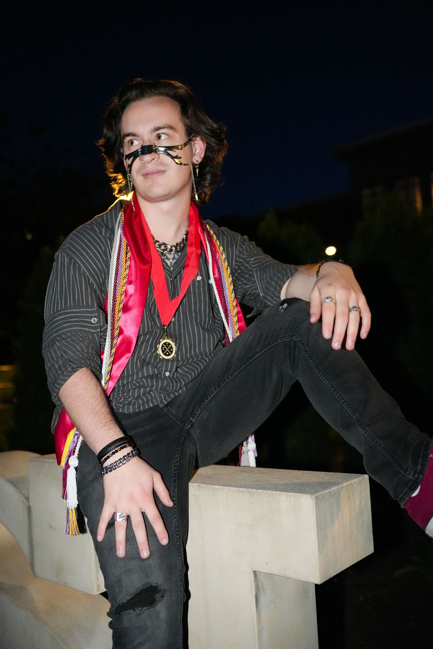 A stylish graduate with curly hair and decorative glasses sits on concrete steps wearing colorful graduation honor cords.