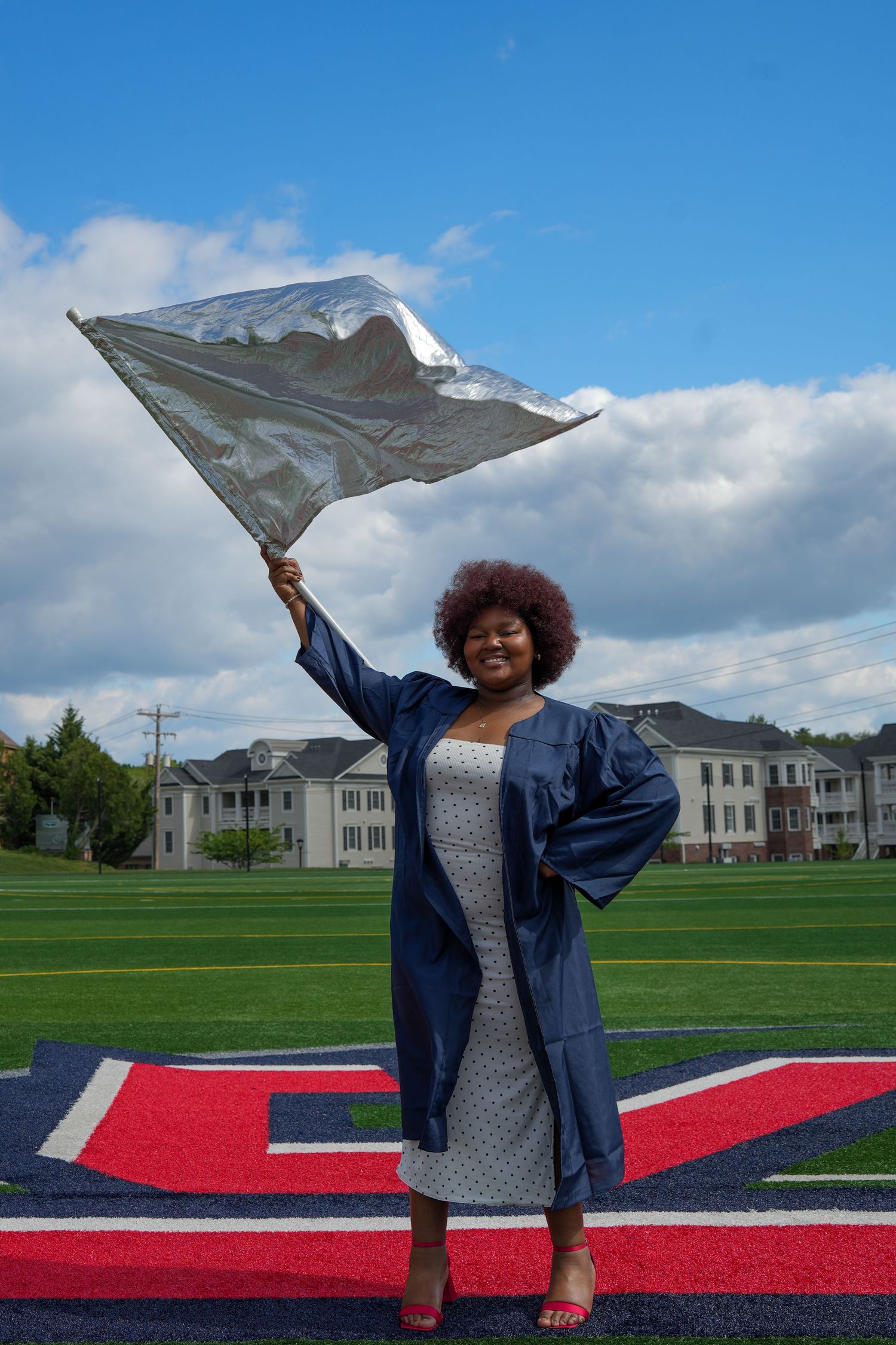 Joyful graduate in blue cap and gown waving silver flag on a green athletic field.