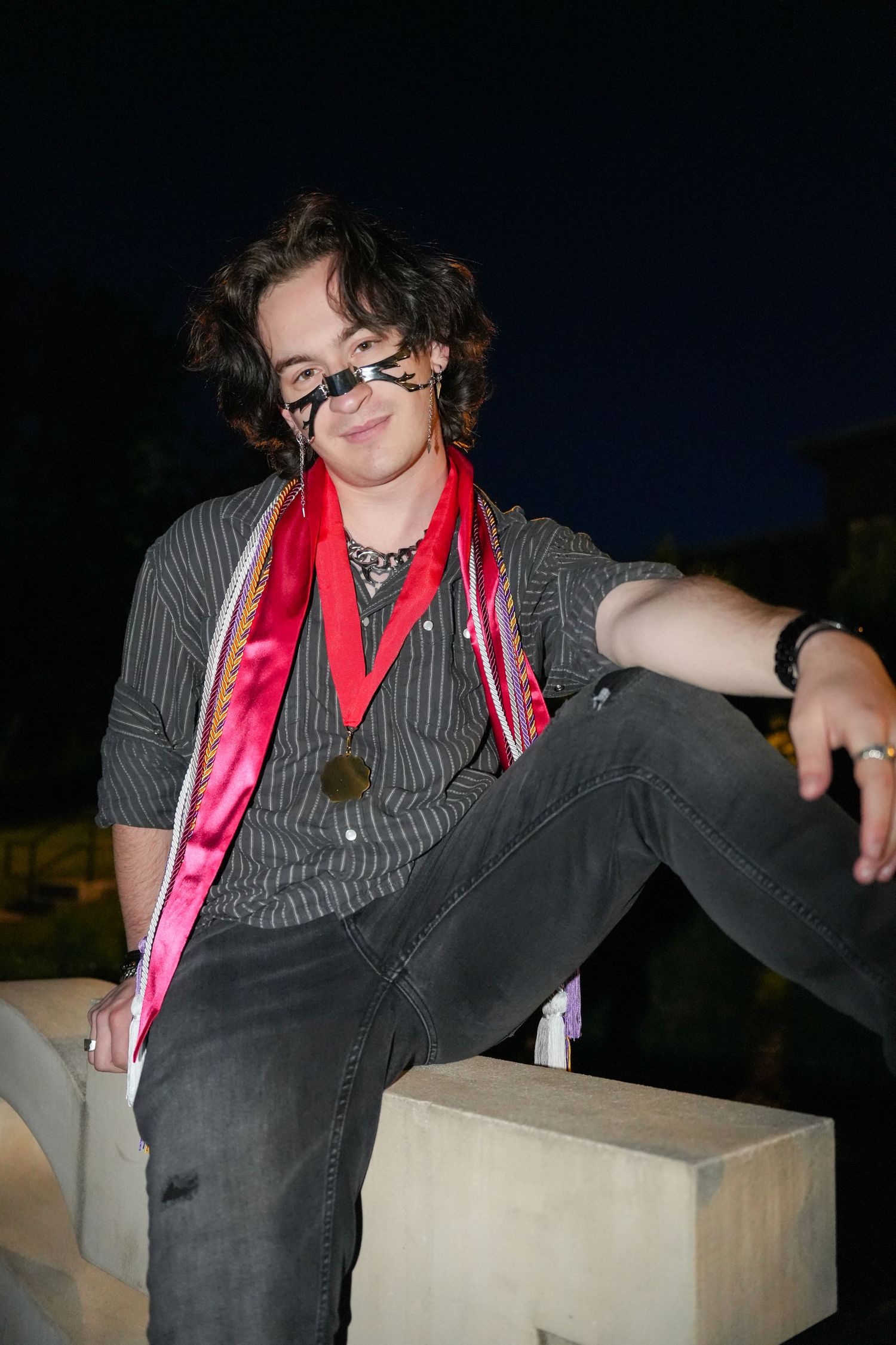 A young person with curly hair and glasses relaxes on concrete steps wearing a red graduation sash at night.