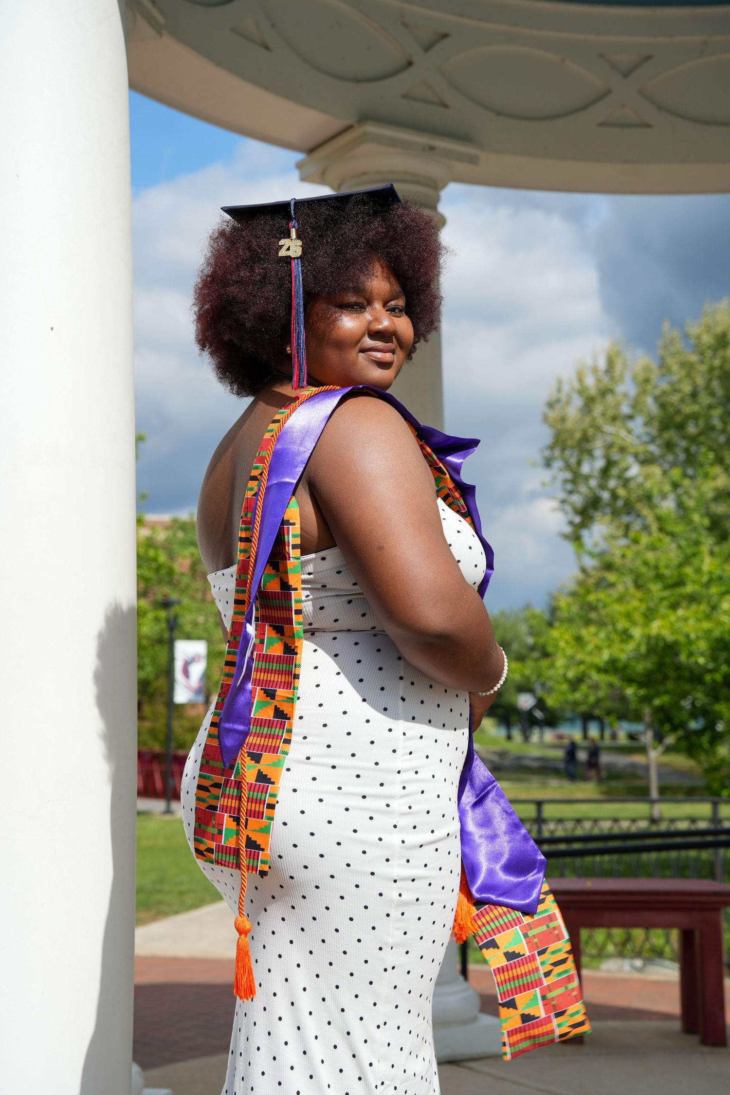 Graduate in white polka dot dress with kente stole and cap posing under outdoor pavilion.