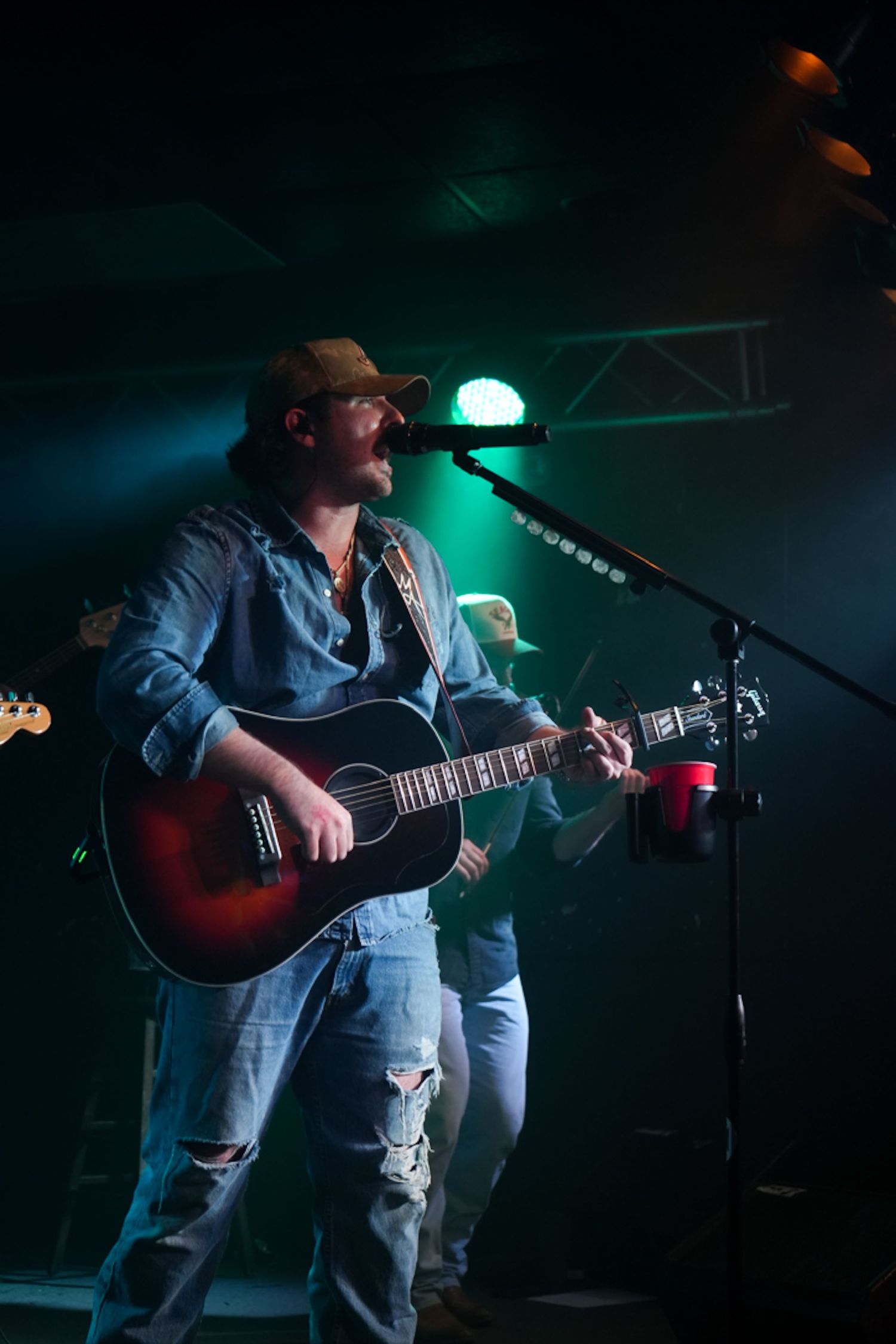 Country musician playing acoustic guitar on stage under green stage lighting.
