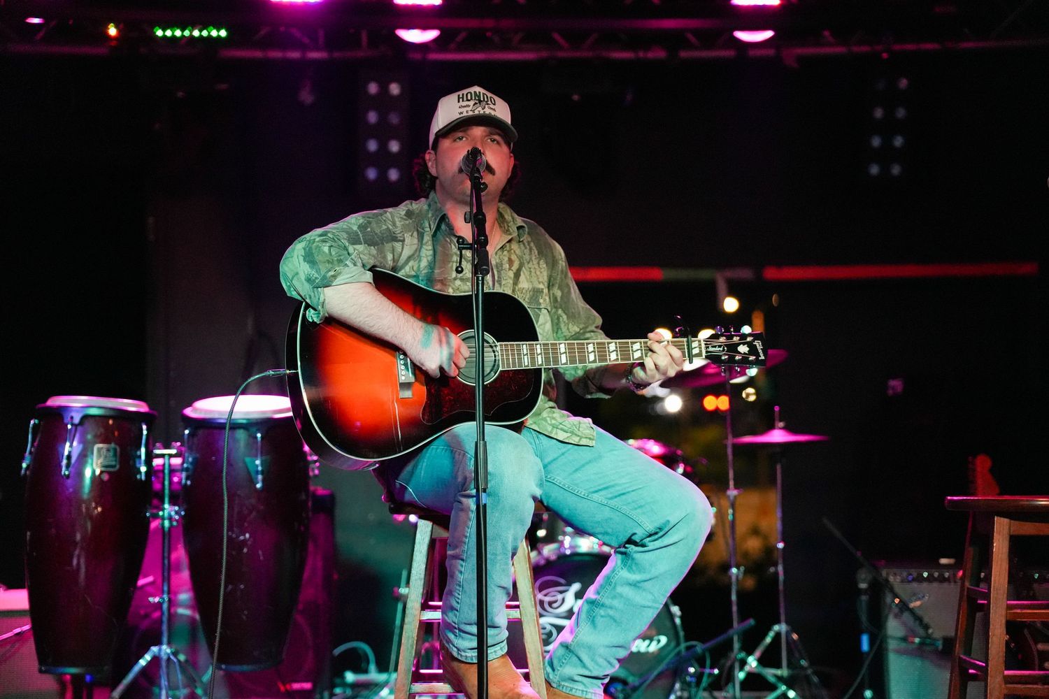 Male musician in cap and floral shirt playing acoustic guitar on colorful stage with conga drums.