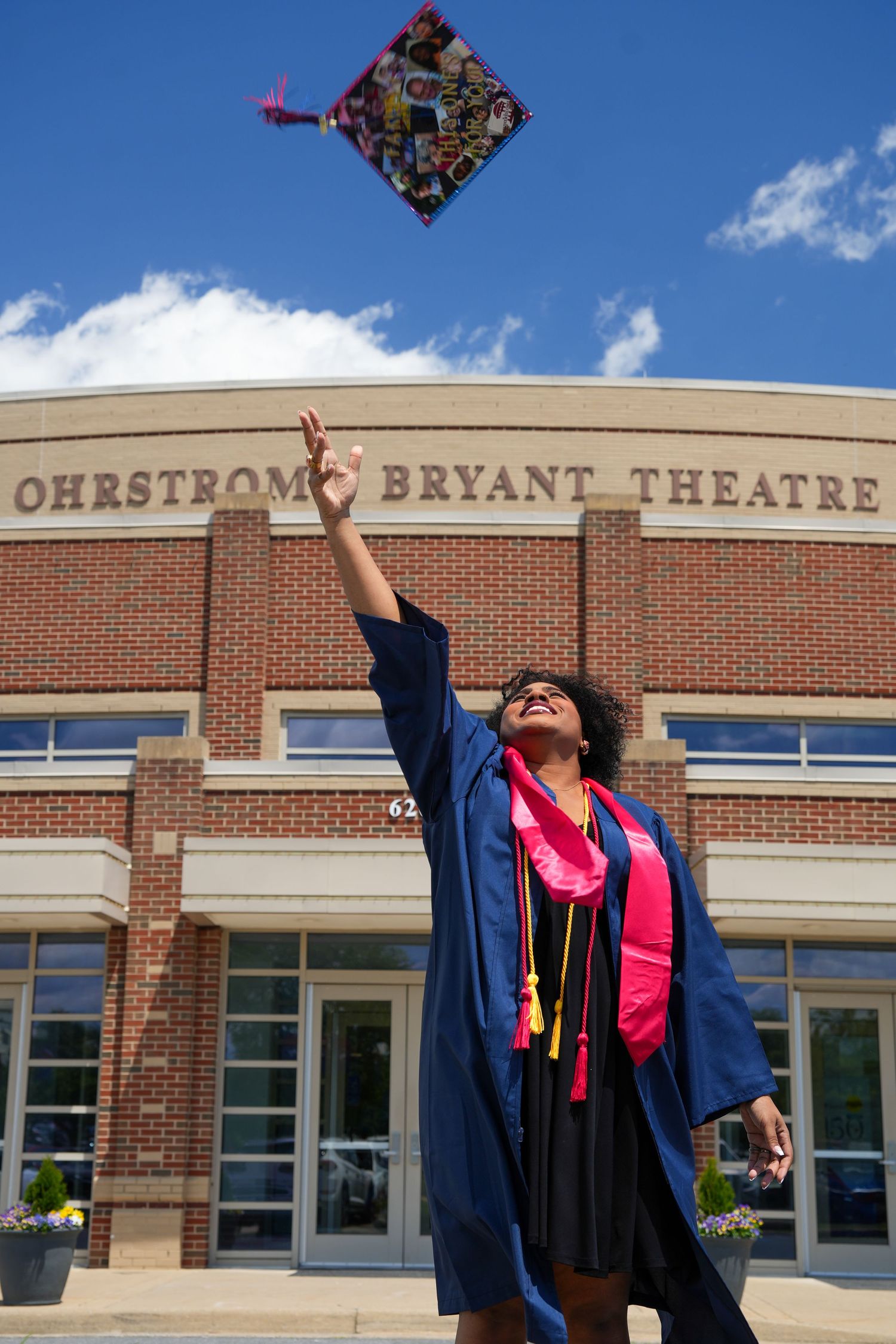 A graduate in blue gown throws decorated mortarboard cap in front of Ohrstrom Bryant Theatre on a sunny day.