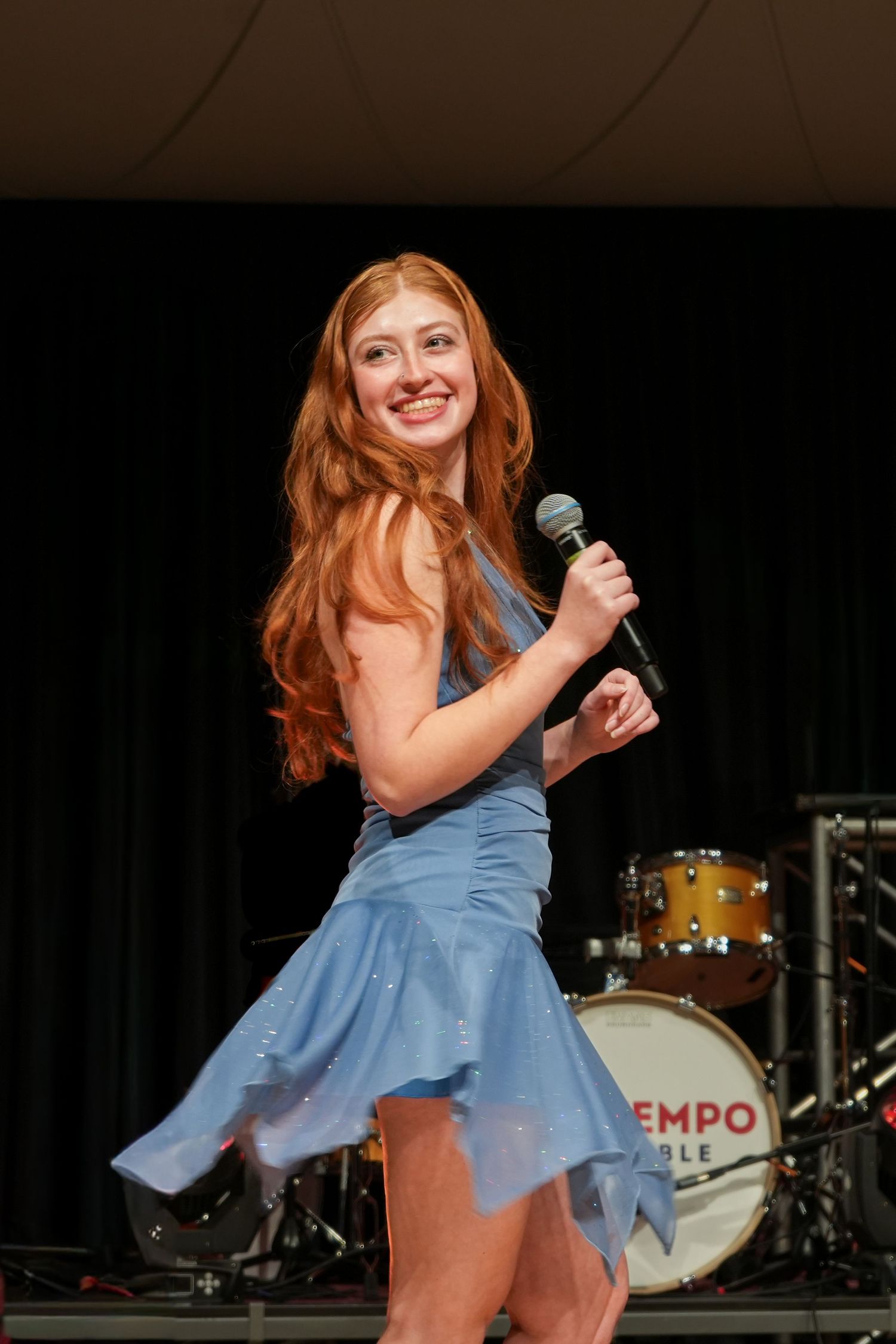 Young redhead woman in blue dress performing on stage with microphone, band drum kit visible behind her.