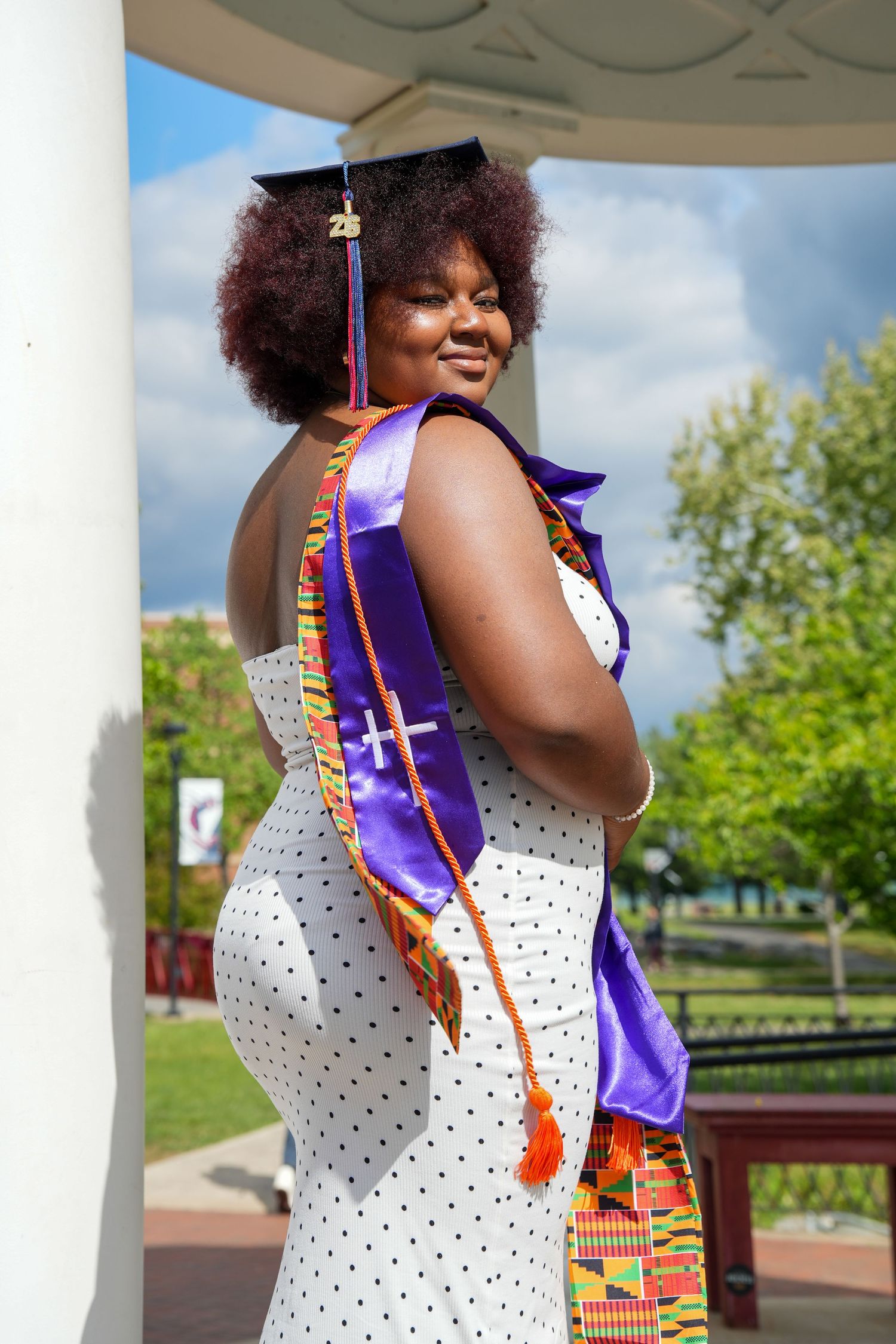 Smiling graduate wearing cap, purple stole, and kente sash posing outdoors on a sunny day.
