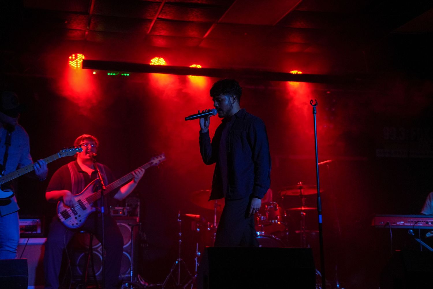 Live band performing on a red-lit stage with guitarist and vocalist holding microphone.