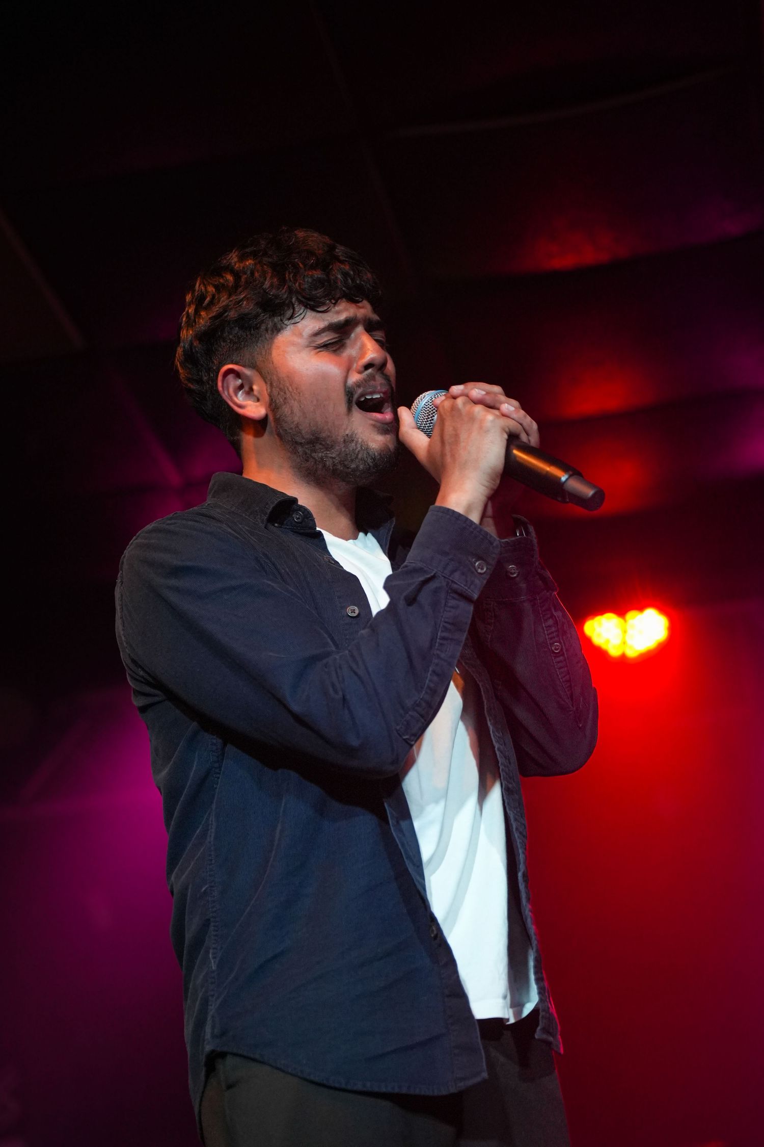 Young male singer performing passionately on stage under red stage lights with microphone.