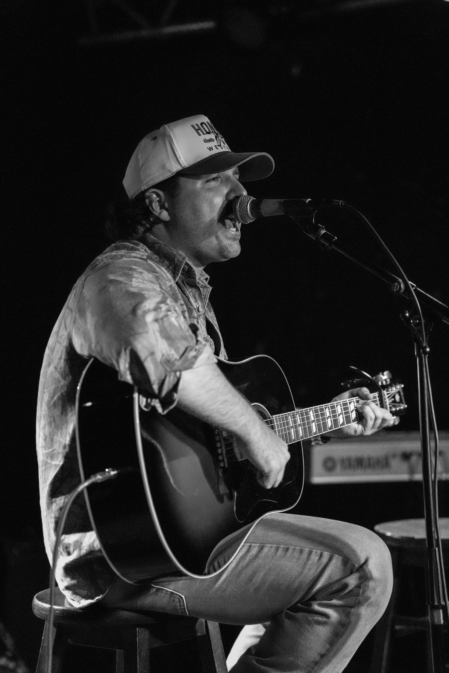 Black and white photo of male guitarist singing at microphone while seated on stage with acoustic guitar.
