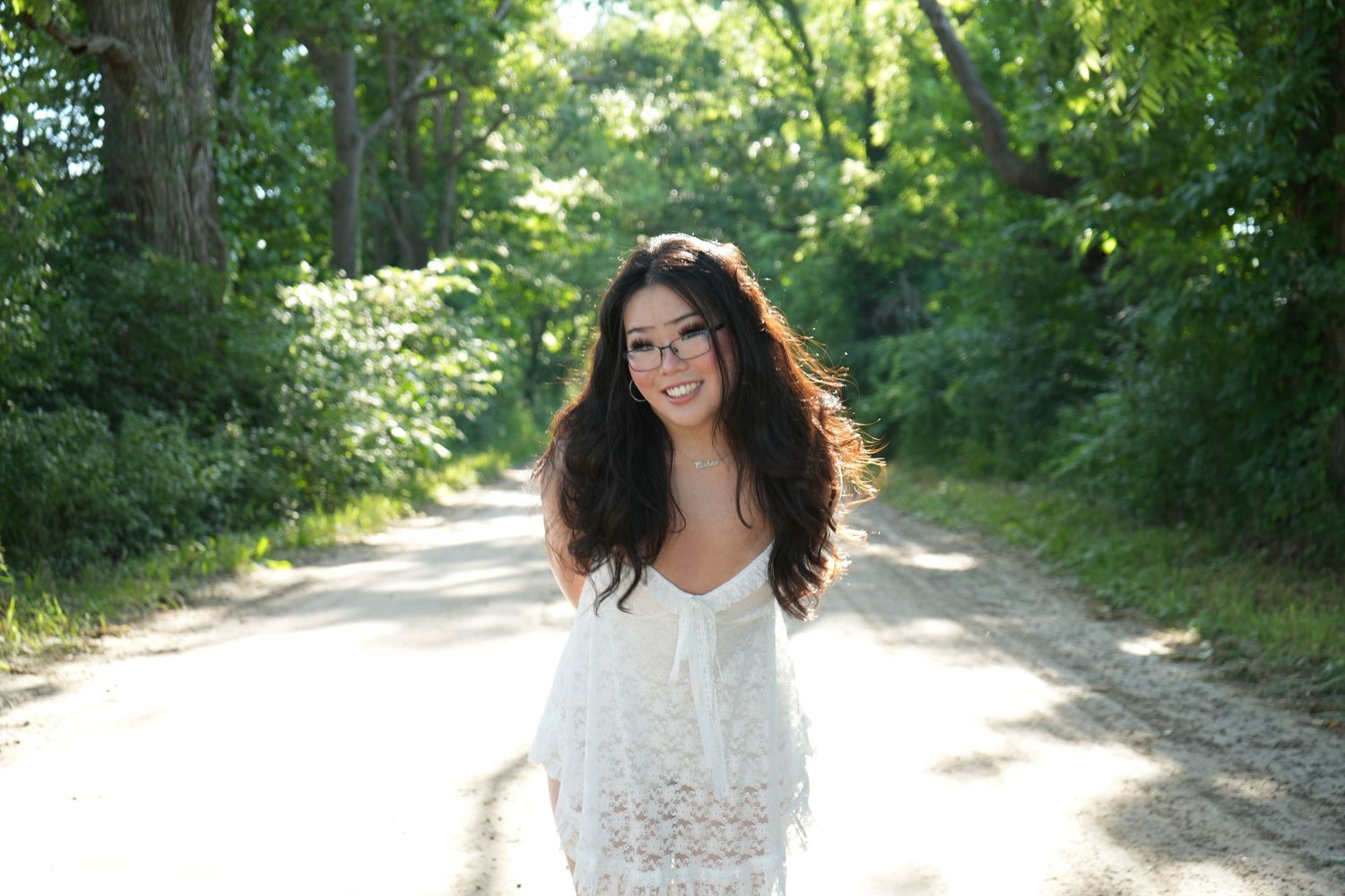 Smiling young woman with long dark hair wearing a white lace dress on a tree-lined country road.