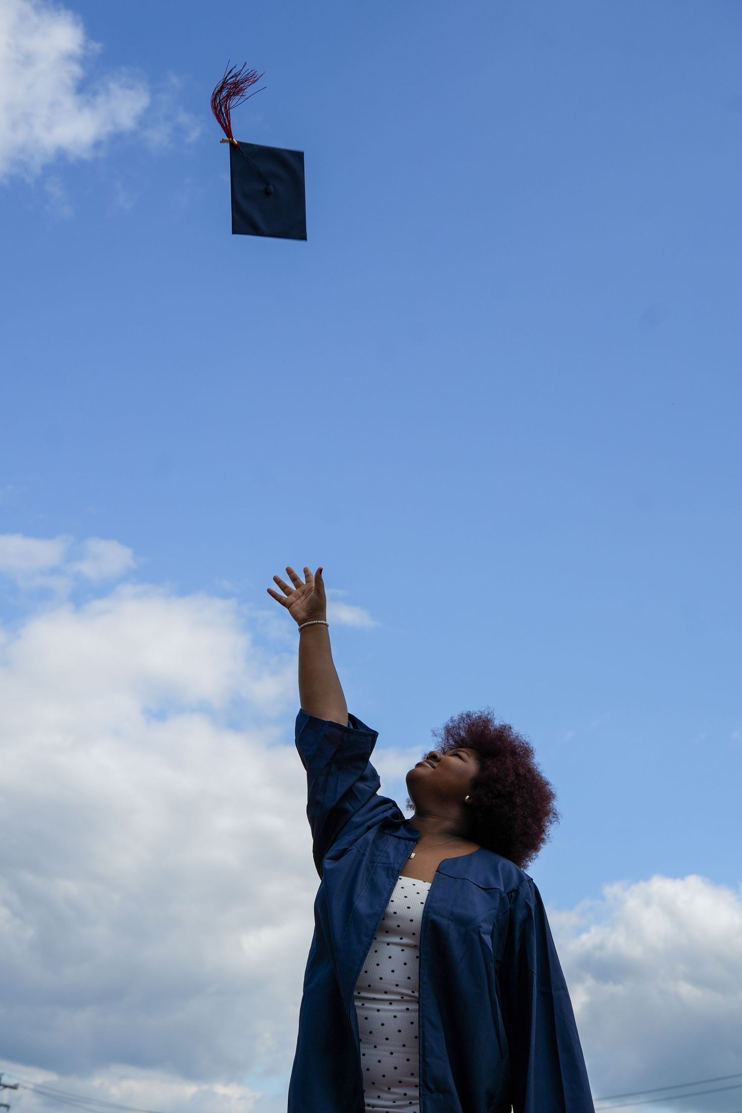 A graduate in blue gown tosses her mortarboard cap into a blue sky, celebrating her academic achievement.
