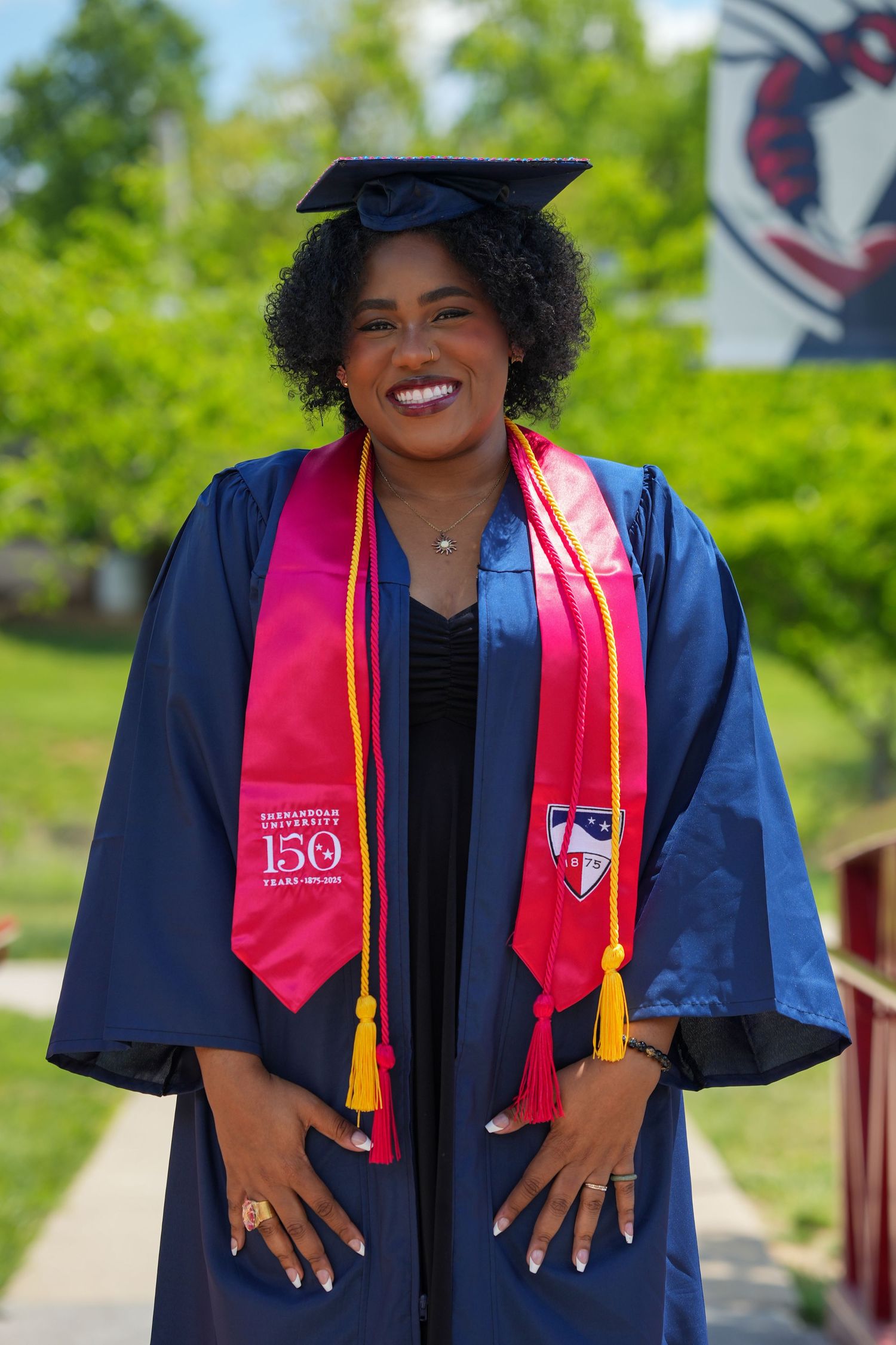 A smiling female graduate in blue cap and gown with pink stole celebrates her 150th anniversary university graduation.