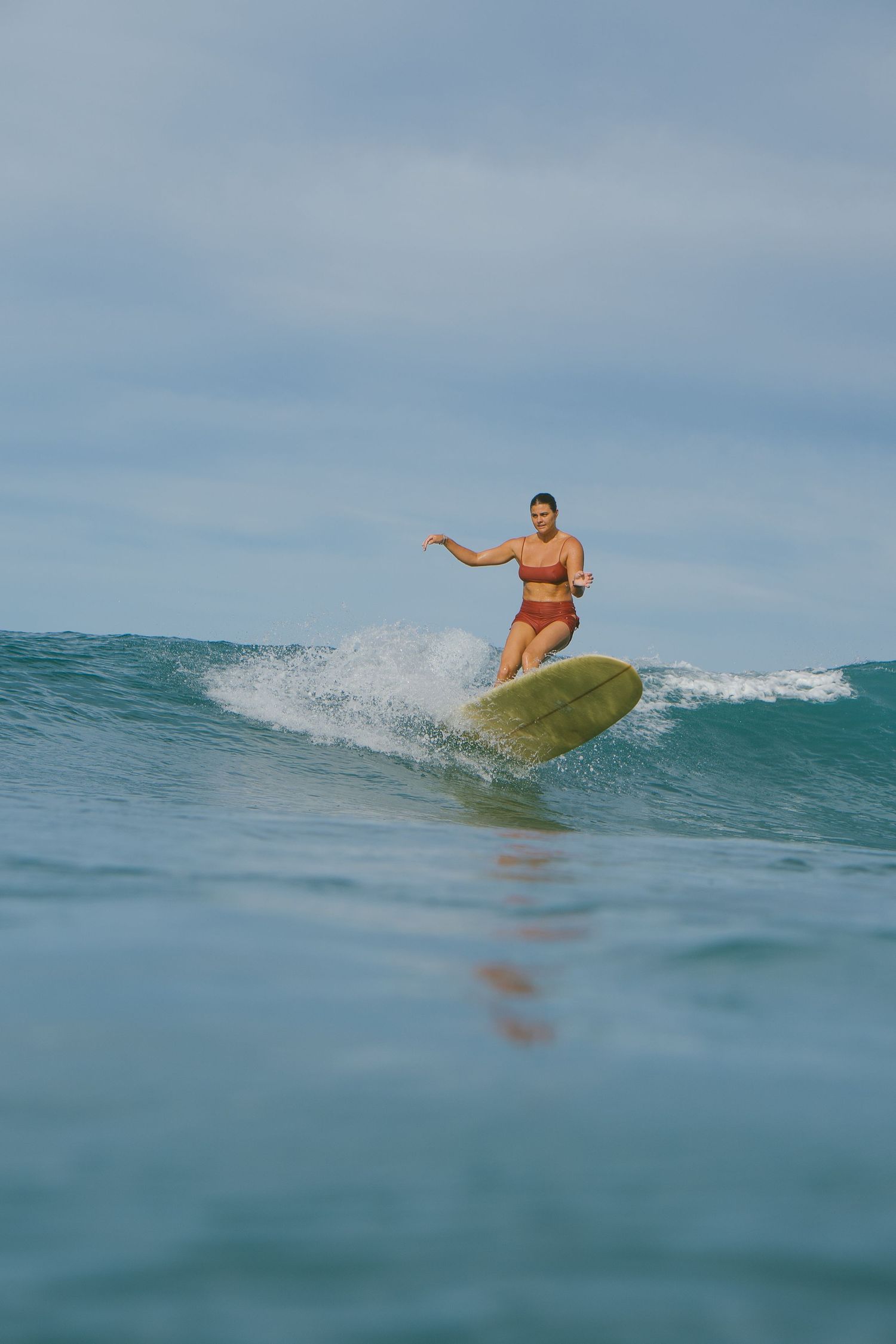 Woman in red bikini surfing a wave on a yellow longboard in clear blue ocean water under cloudy skies.