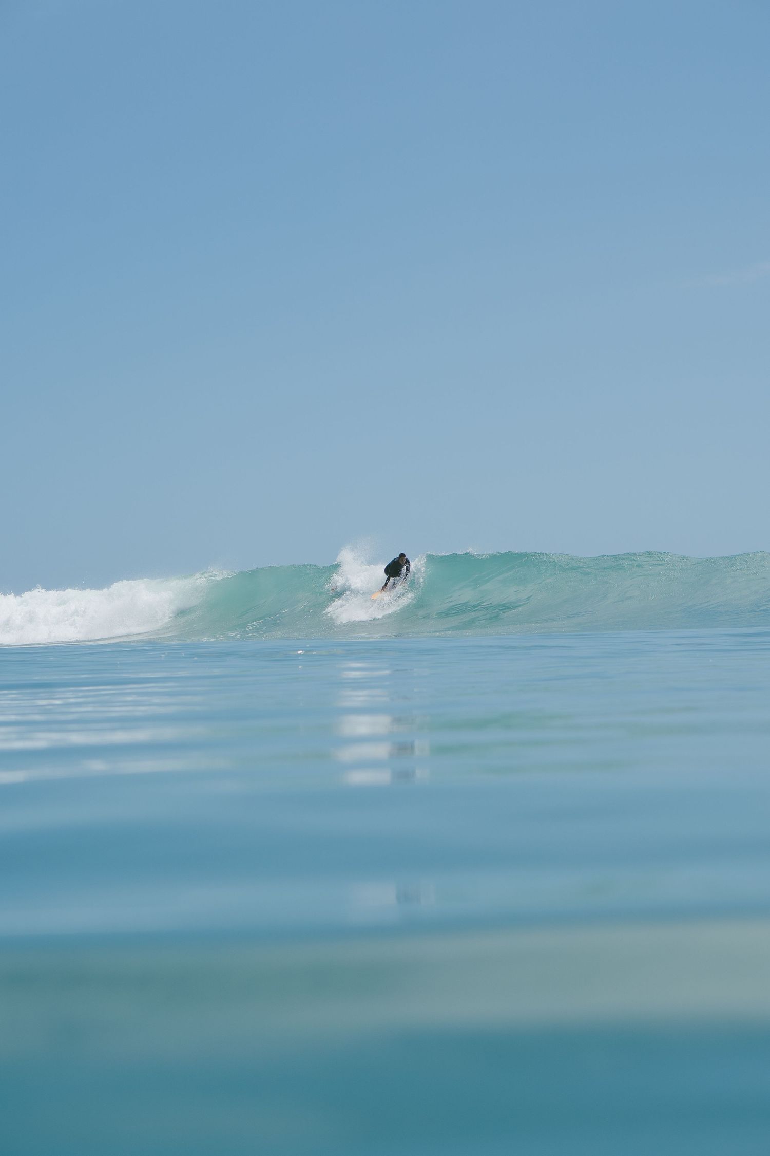 Long-distance shot of a surfer riding a pristine turquoise wave against a clear blue sky.