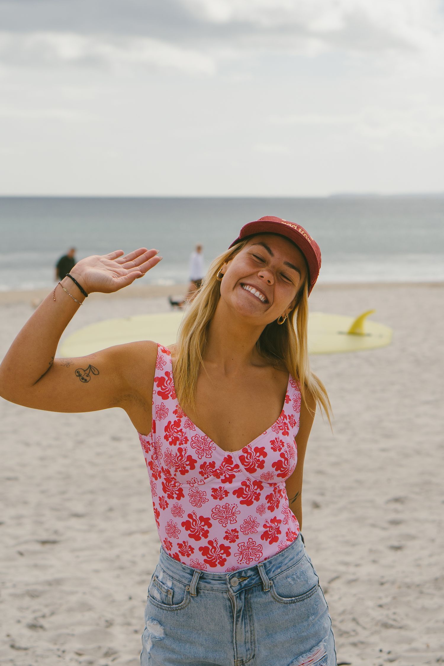 Person in floral swimsuit and denim shorts poses joyfully on sunny beach.