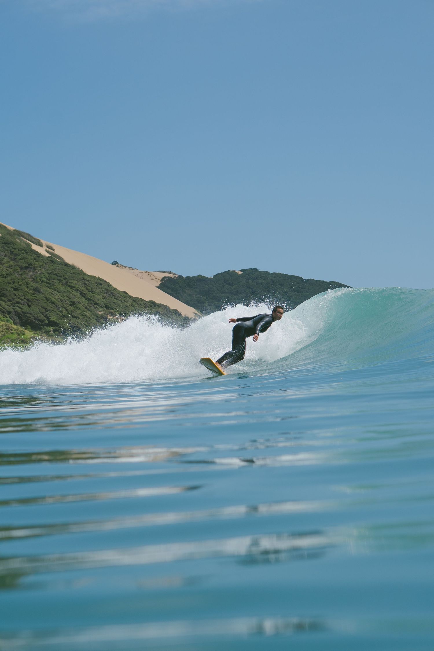 Surfer catches air off breaking wave with sand dunes and green hills in background.