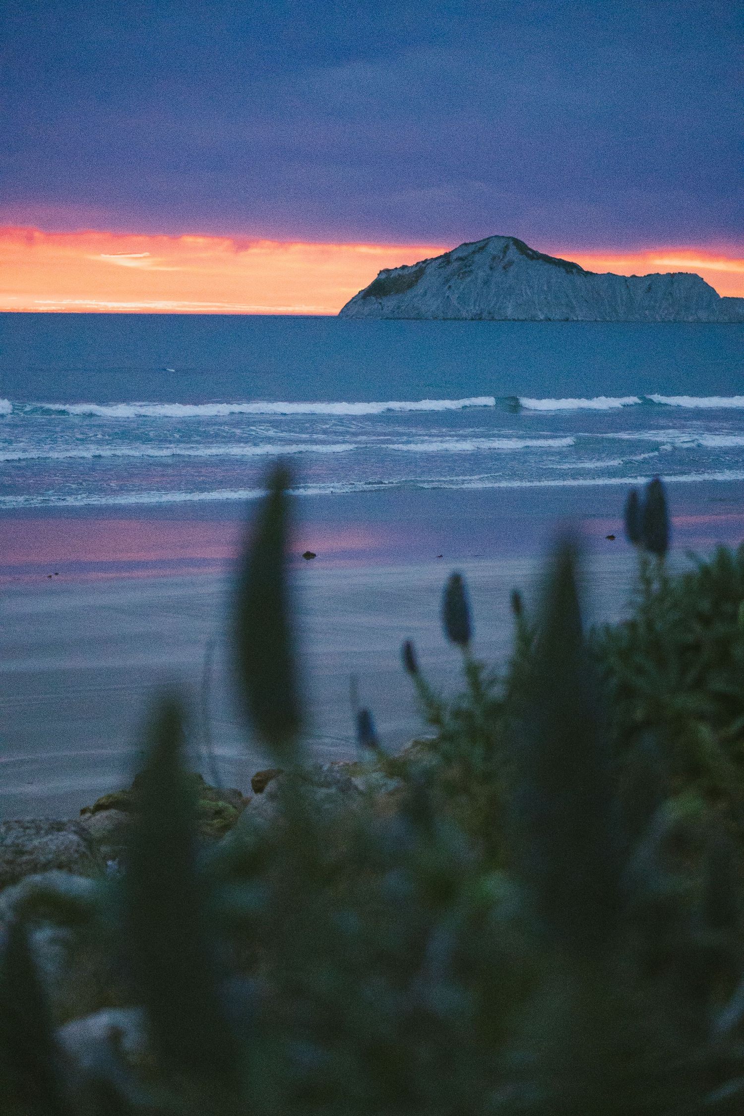Scenic coastal sunset view with mountains silhouetted against an orange and purple sky.