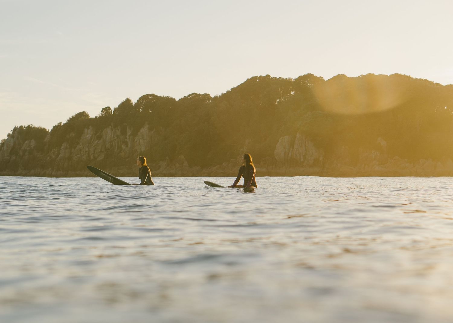 Golden sunset over ocean waters with distant surfers visible near rocky coastline.