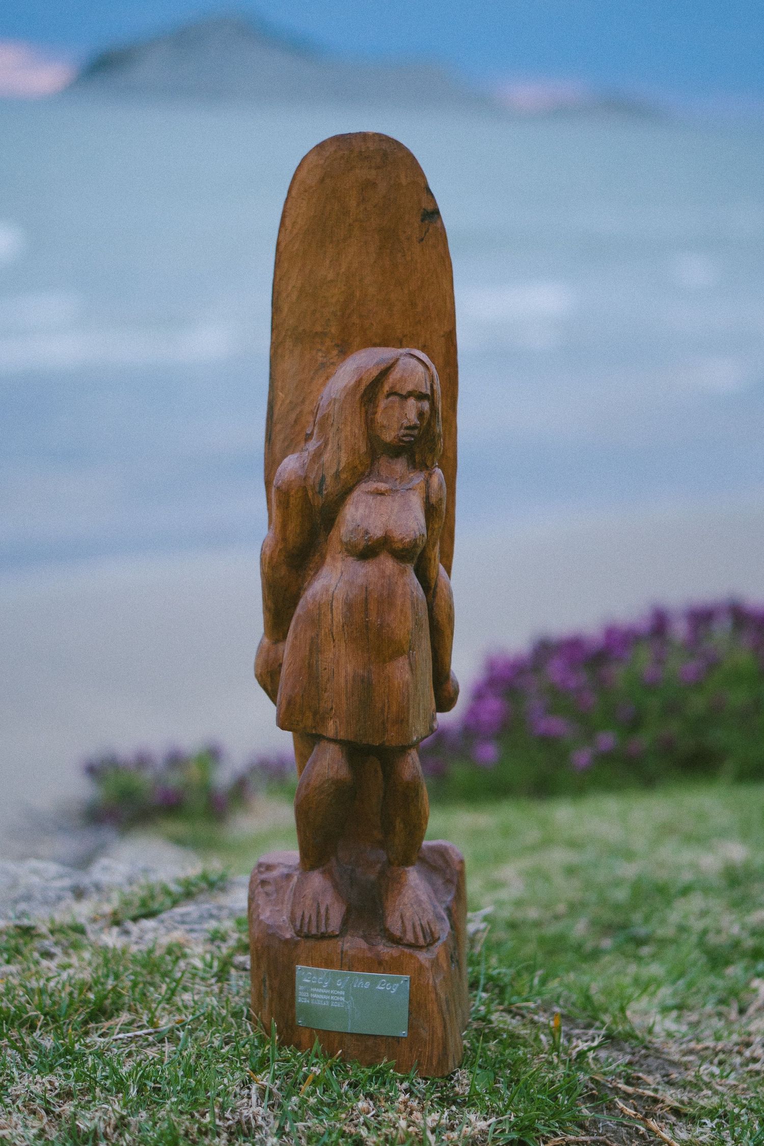 Wooden carved statue or sculpture displayed on grass with purple flowers in the background.