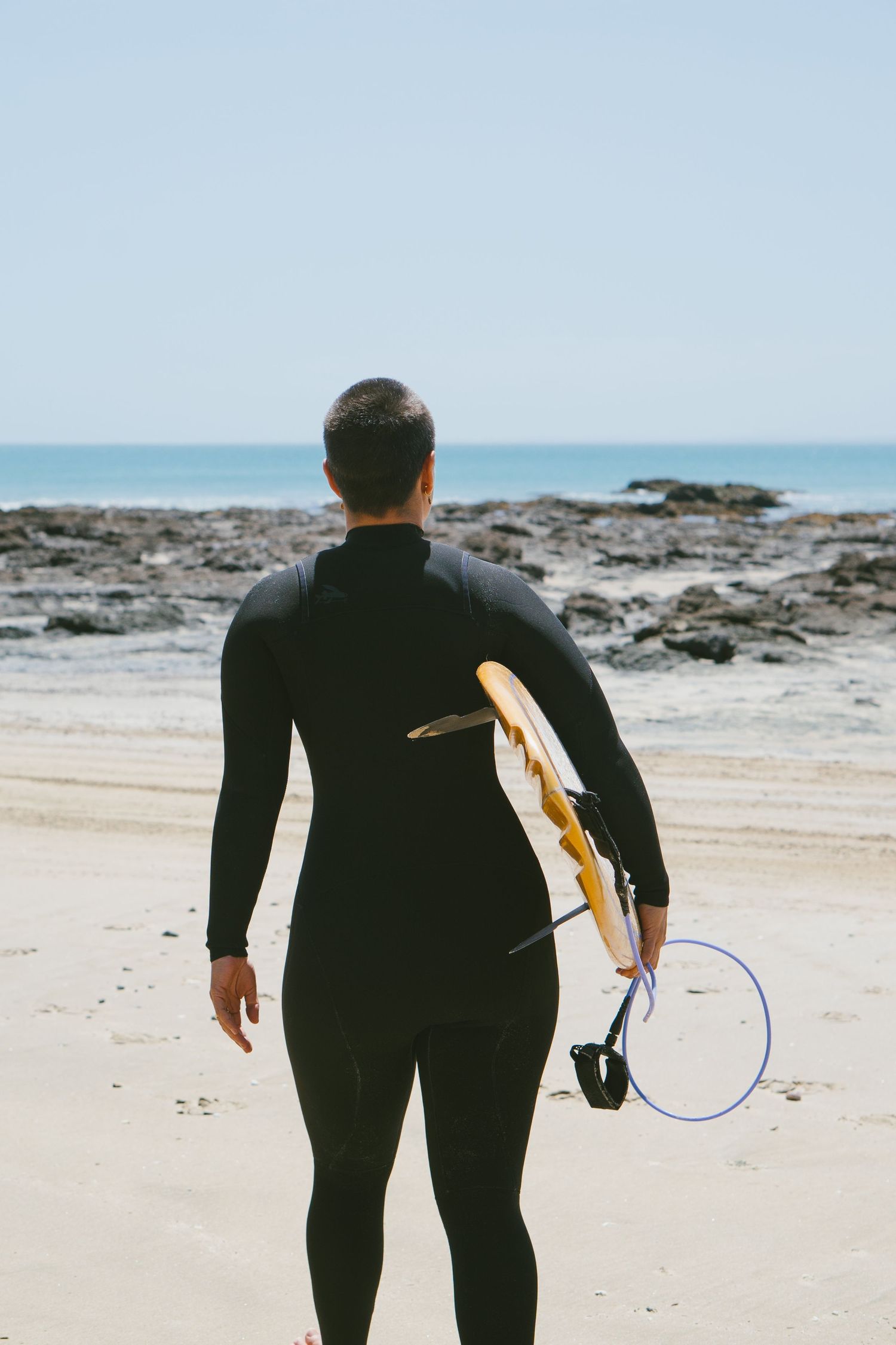A surfer in a black wetsuit carries a surfboard across a rocky beach on a sunny day.