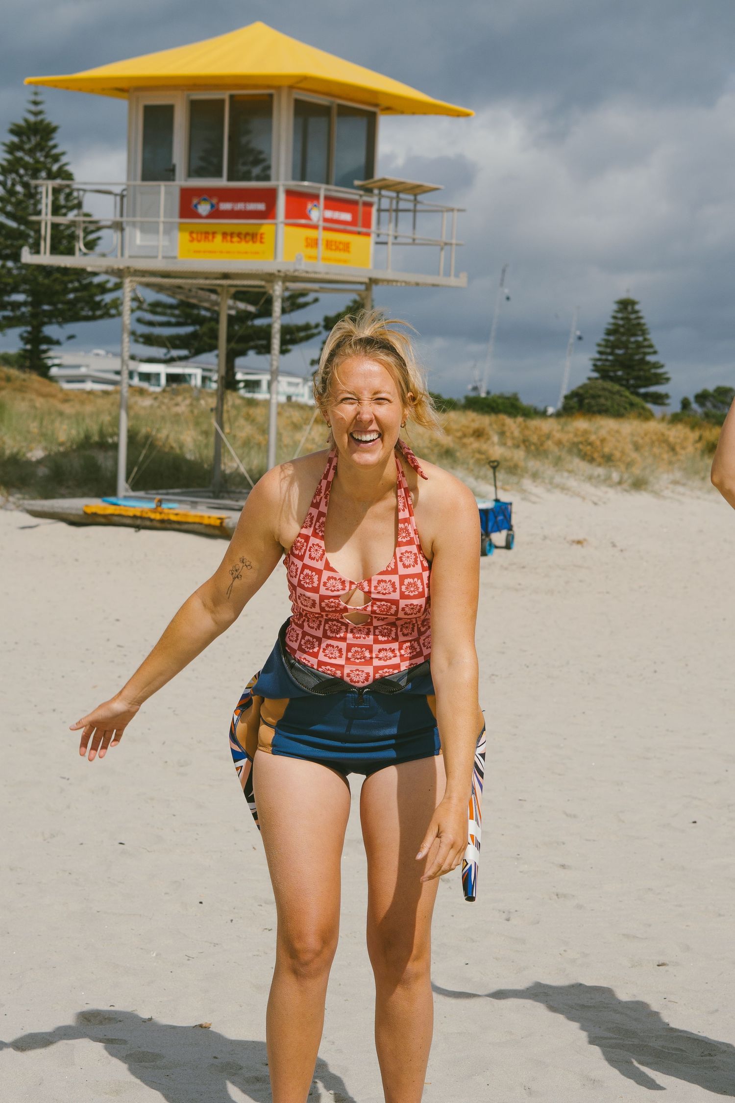 Person in red swimwear poses playfully on beach with yellow lifeguard tower in background.