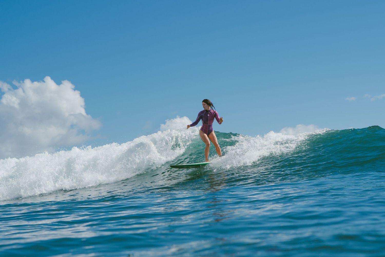 Surfer gracefully rides ocean wave in purple wetsuit against blue sky.