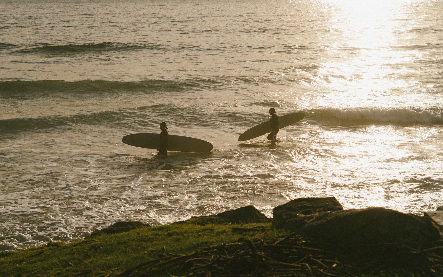 Silhouettes of surfers walking along rocky coast at sunset.
