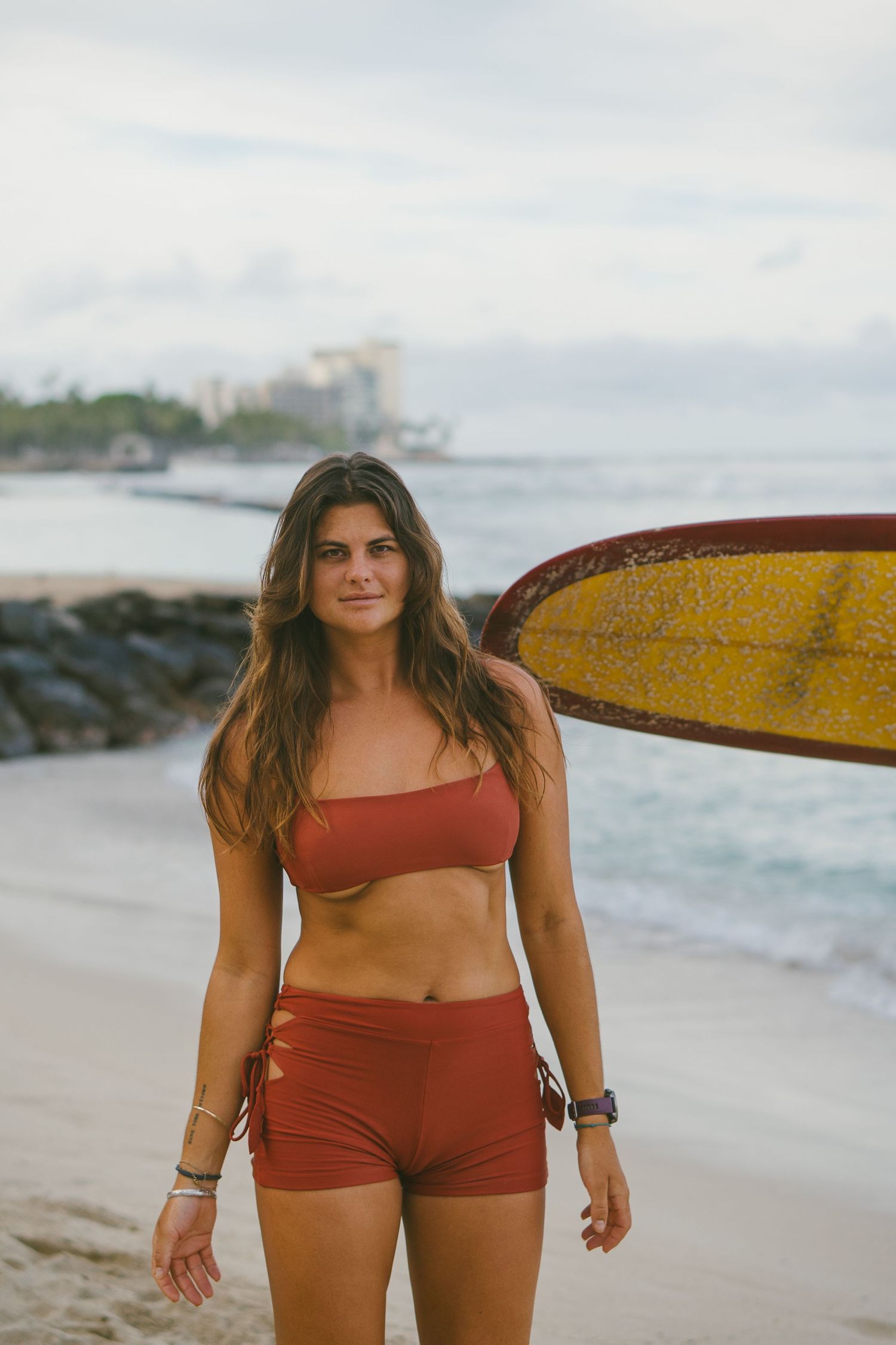 A young woman in a rust-colored bikini set carries a yellow surfboard on a sandy beach.