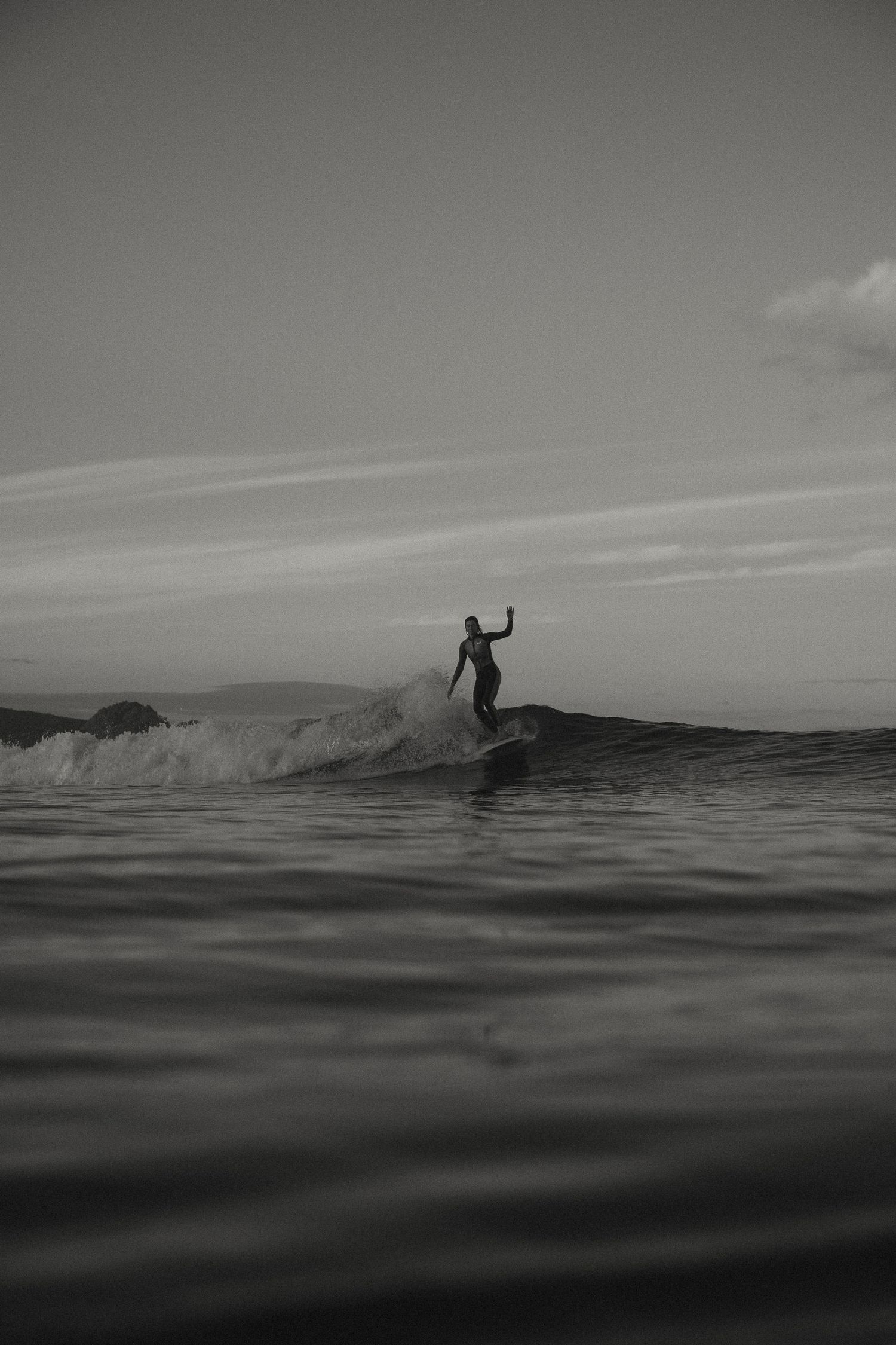 Distant surfer silhouettes catching waves on a moody ocean during sunset.
