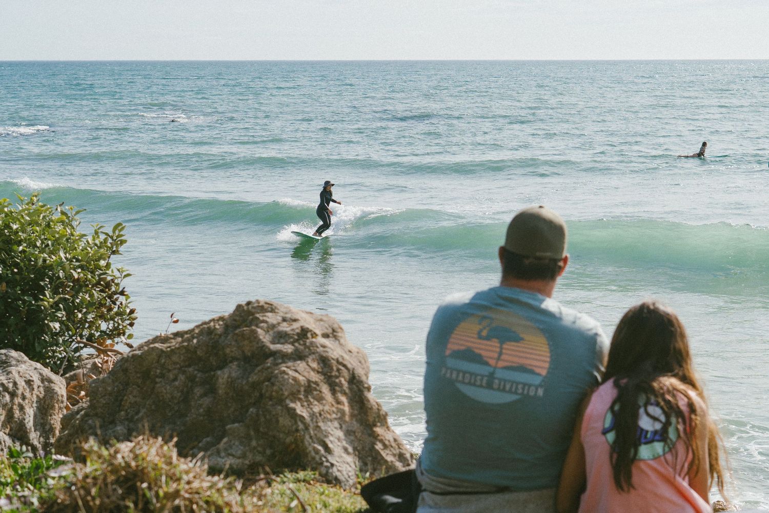 Spectators watching surfers ride waves from rocky coastal viewpoint.