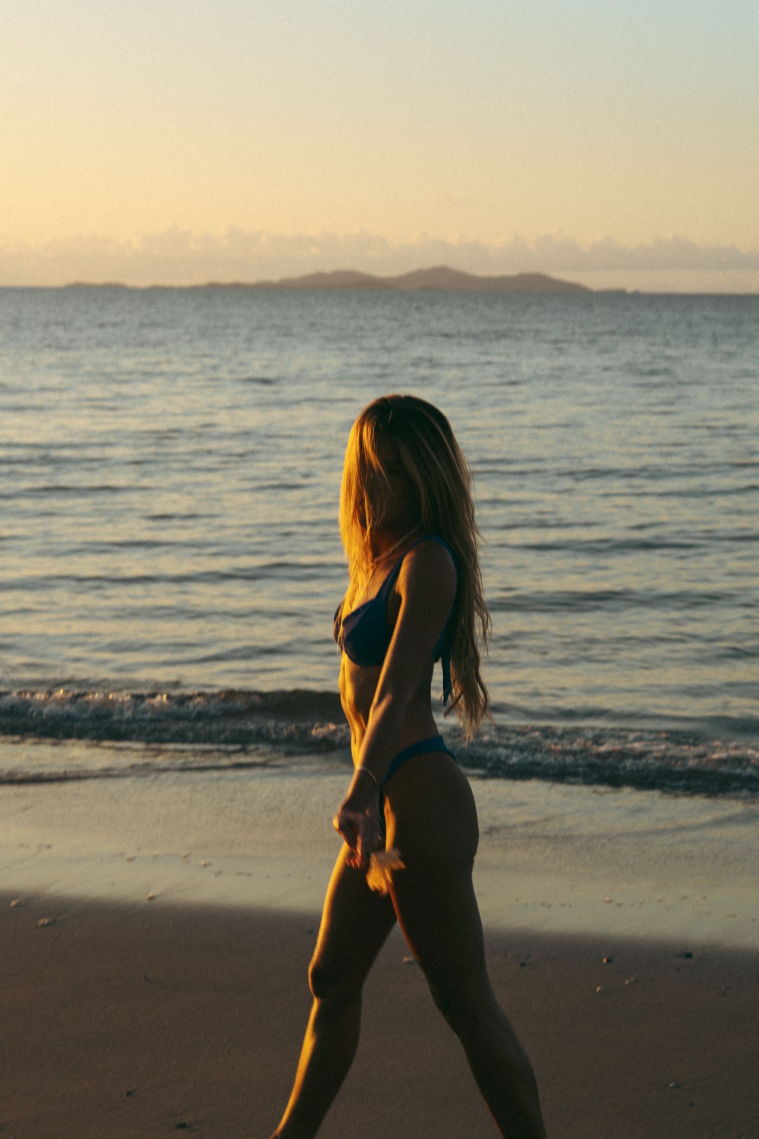 Silhouetted figure walks along a beach at sunset with ocean waves and mountains in the background.