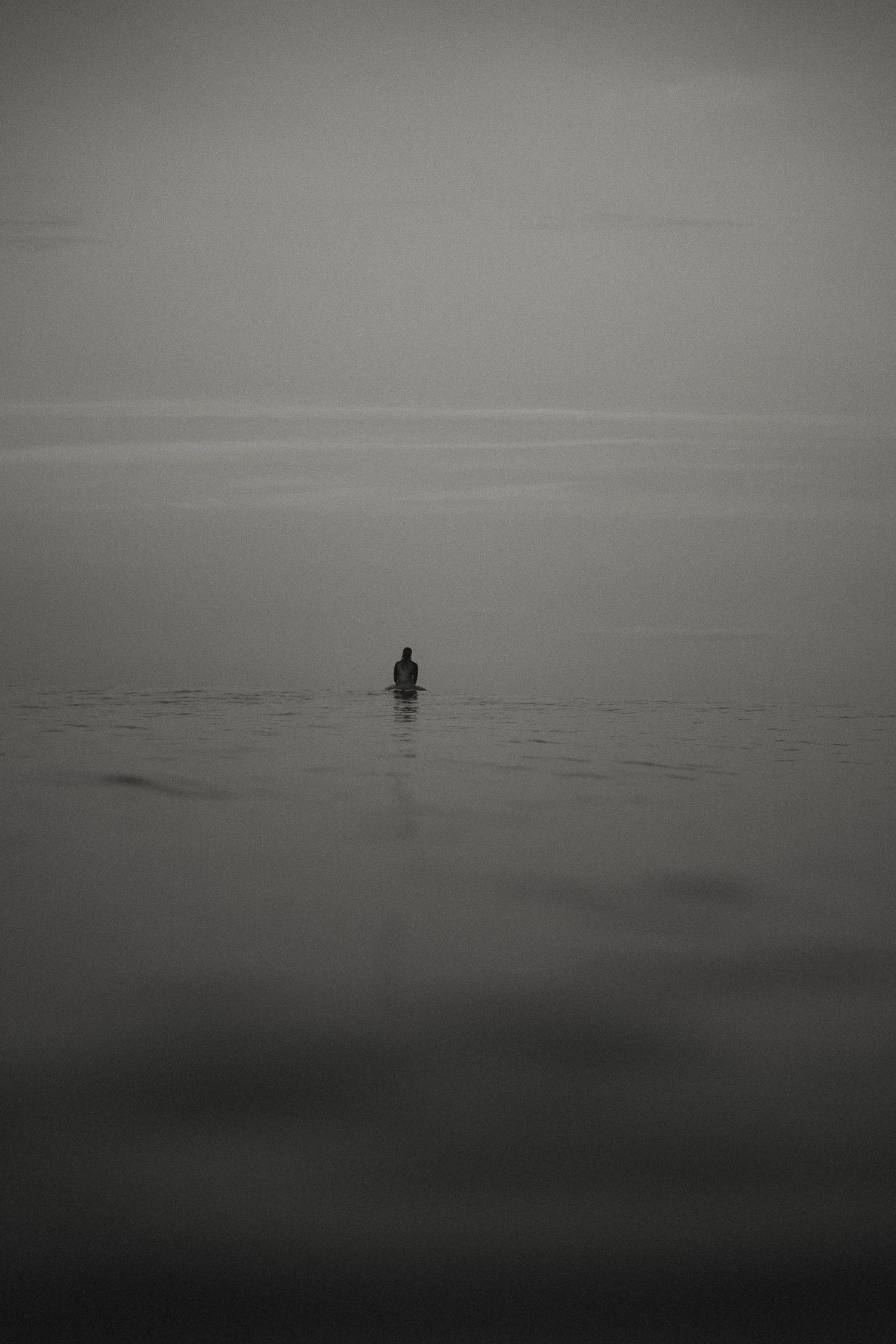 A lone surfer sits on their board in calm misty waters during an overcast day.