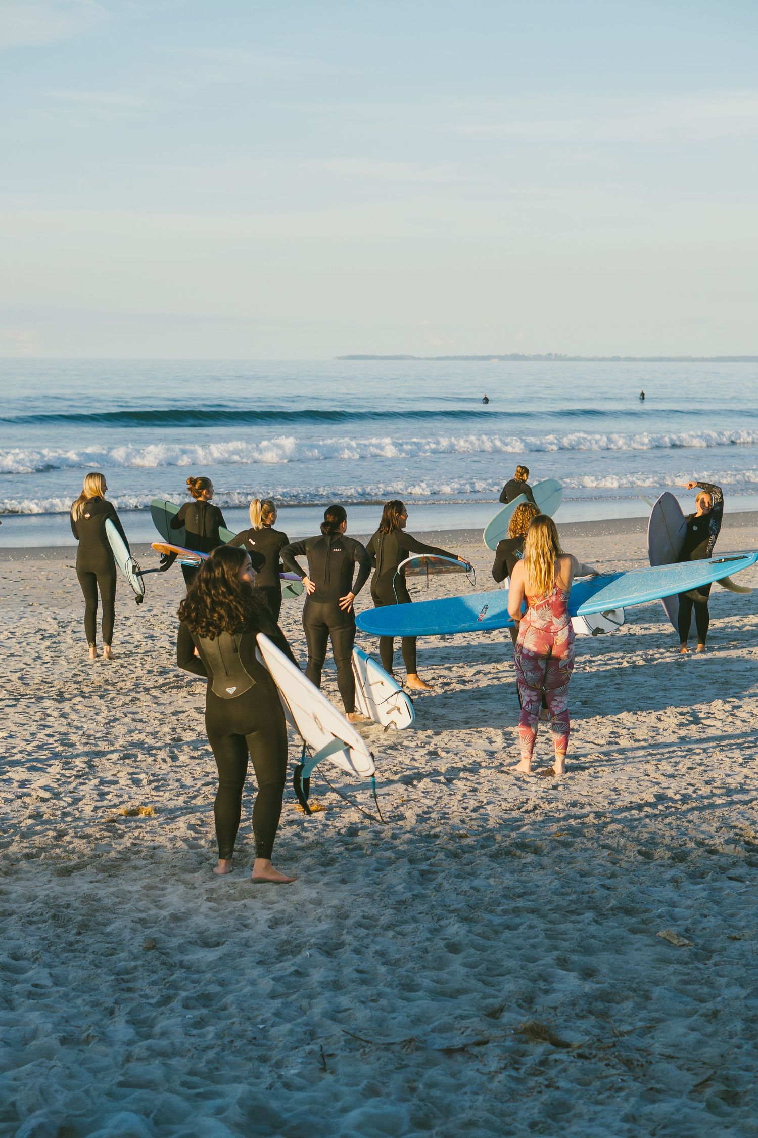 Group of surfers in wetsuits carrying surfboards toward ocean waves on a sunny sandy beach.