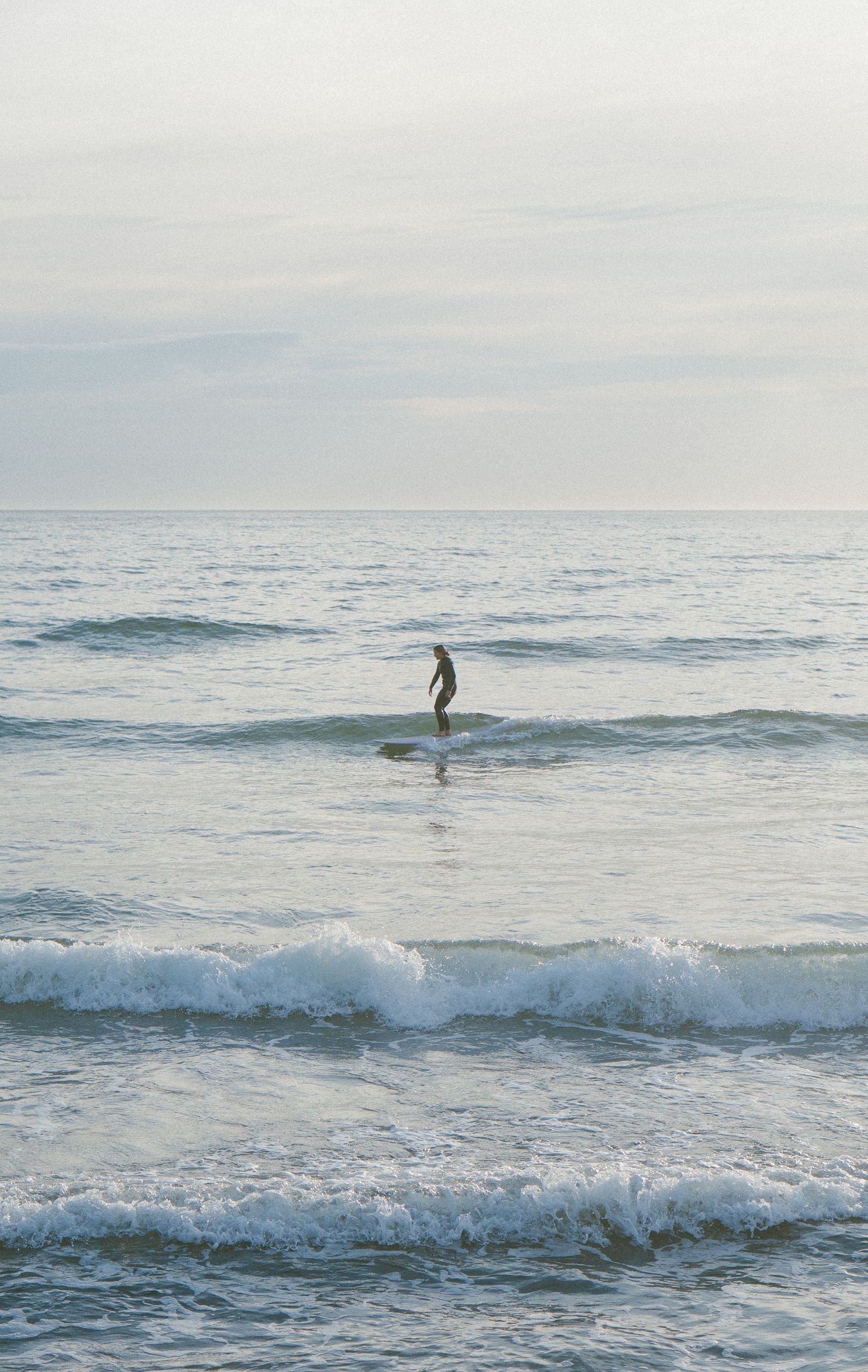 Lone surfer rides waves in misty morning ocean under cloudy sky.