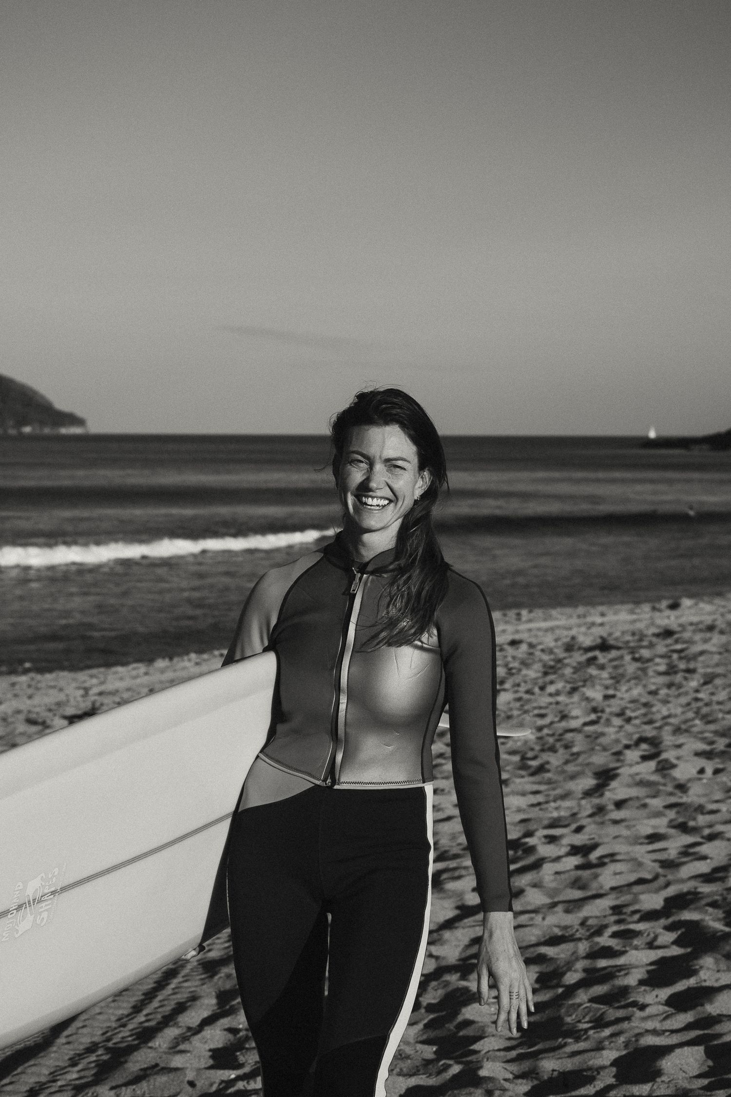 Surfer in black wetsuit poses with surfboard on beach in black and white photography.