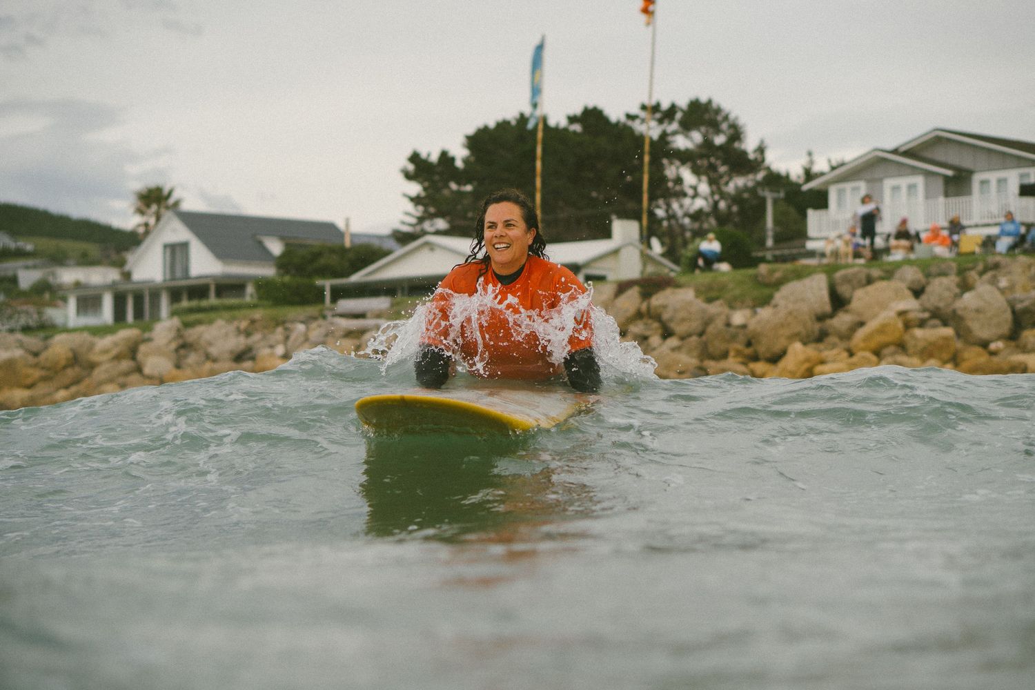 Surfer riding waves near a rocky coastline with houses visible in the background.
