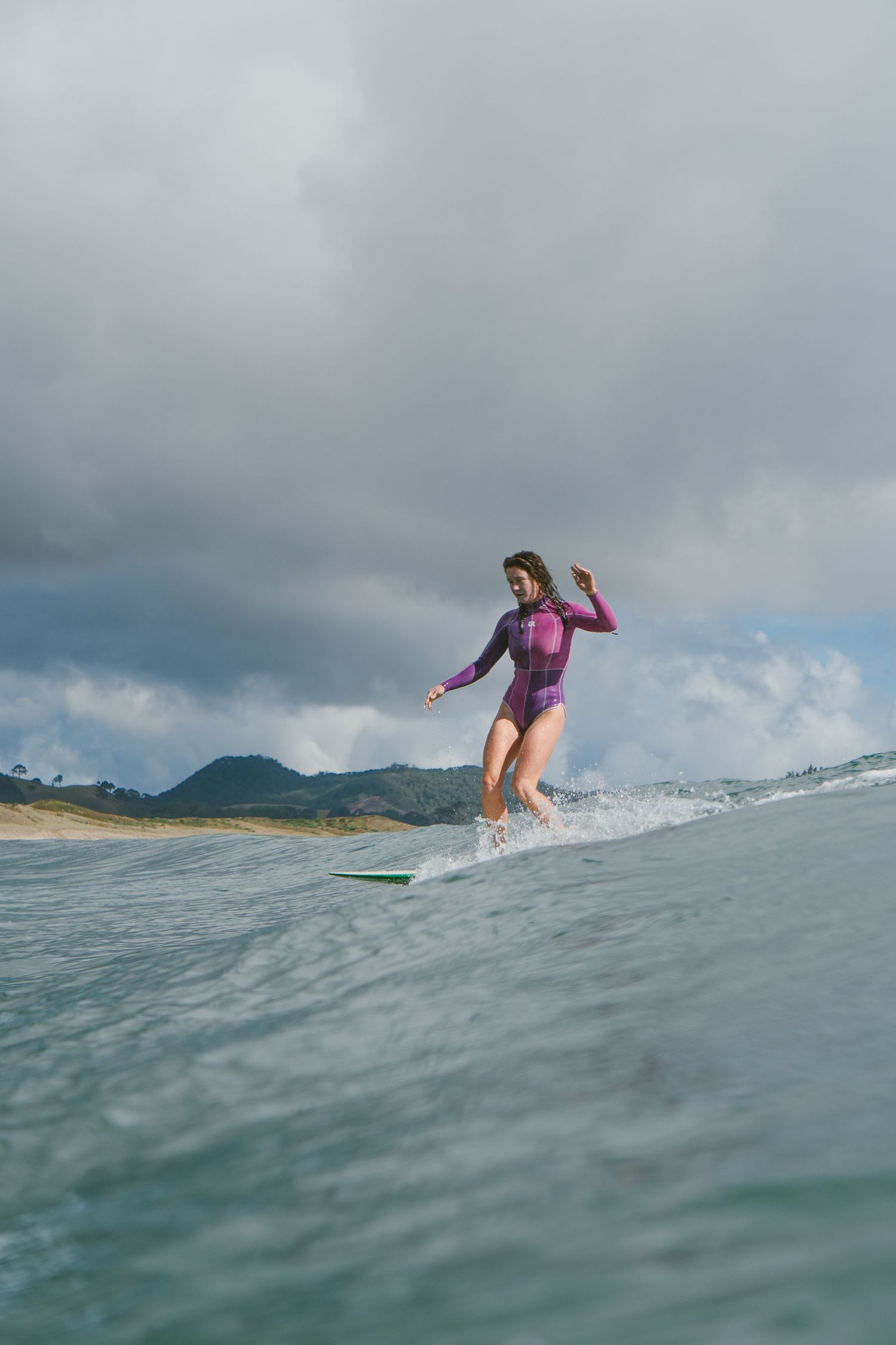 Surfer in purple wetsuit rides ocean wave against dramatic cloudy sky and mountain backdrop.