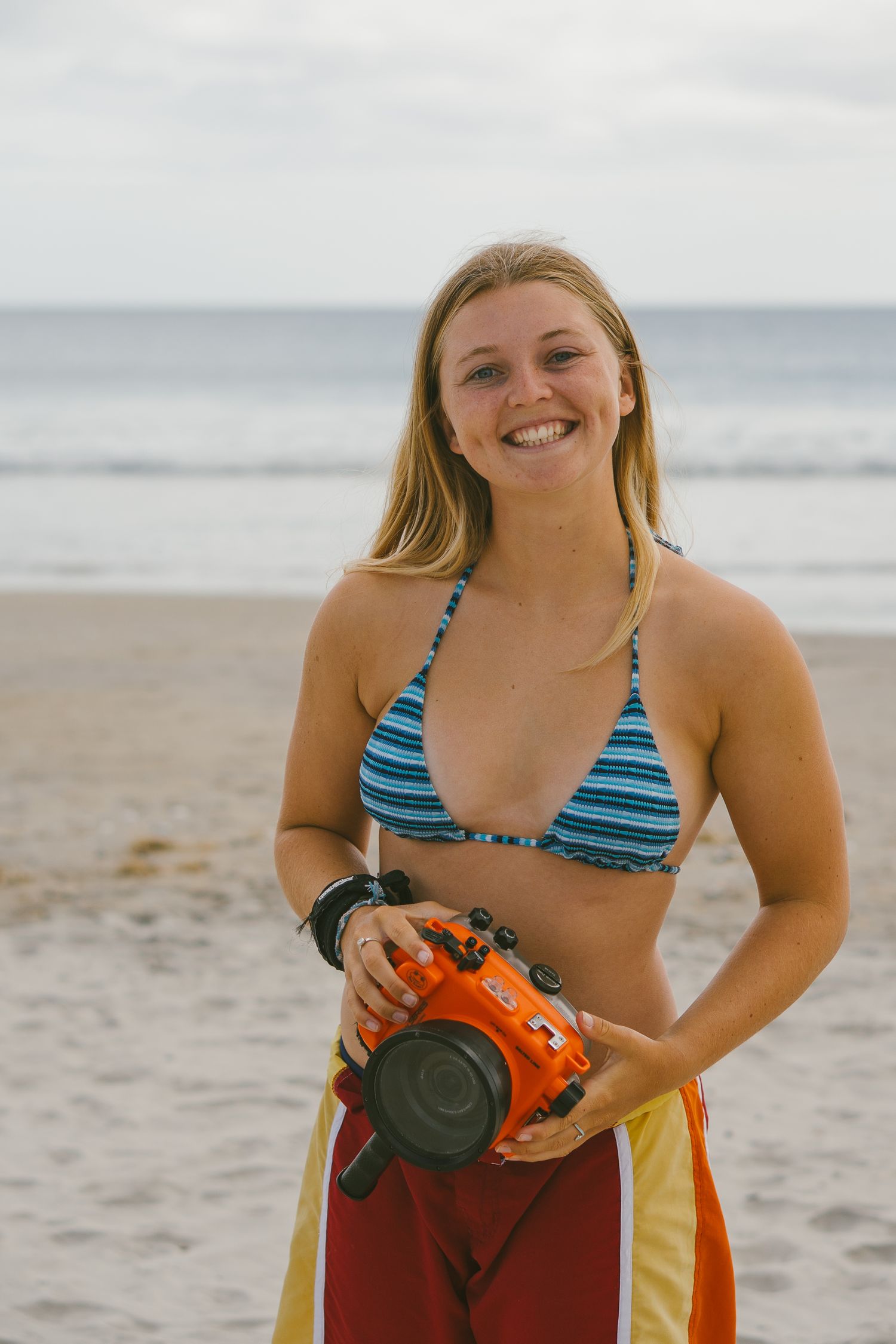 Person in striped swimwear holding camera gear on sandy beach.